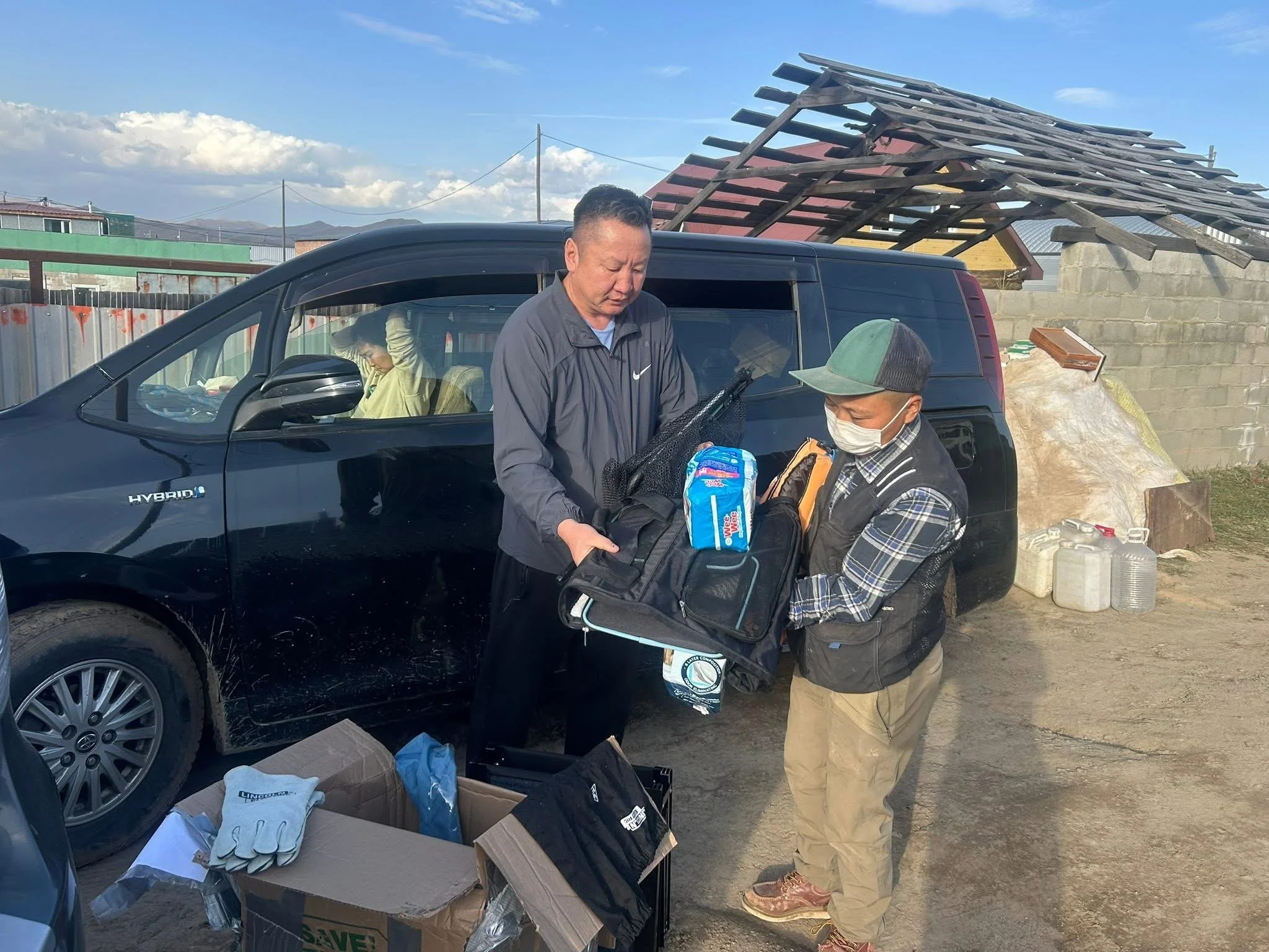 Two men are unloading gear from a black hybrid vehicle outdoors, with one man wearing a face mask and a baseball cap while the other is dressed in a gray jacket. The background includes a partially covered outdoor structure, miscellaneous containers, and a cloudy sky.