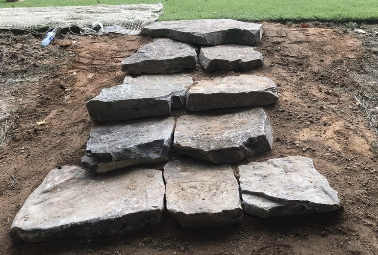 Unfinished stone pathway with large, flat stones placed in a row on dirt, with some grass and construction materials in the background.
