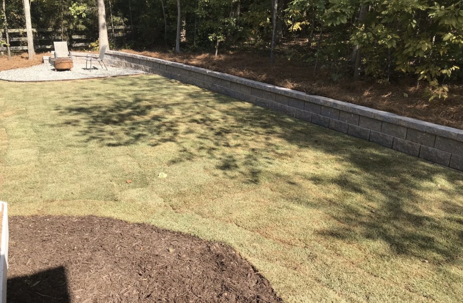 A backyard with a well-maintained lawn, a curved garden bed with mulch in the foreground, a stone retaining wall along the back, trees and bushes, and outdoor seating including two chairs and a fire pit.