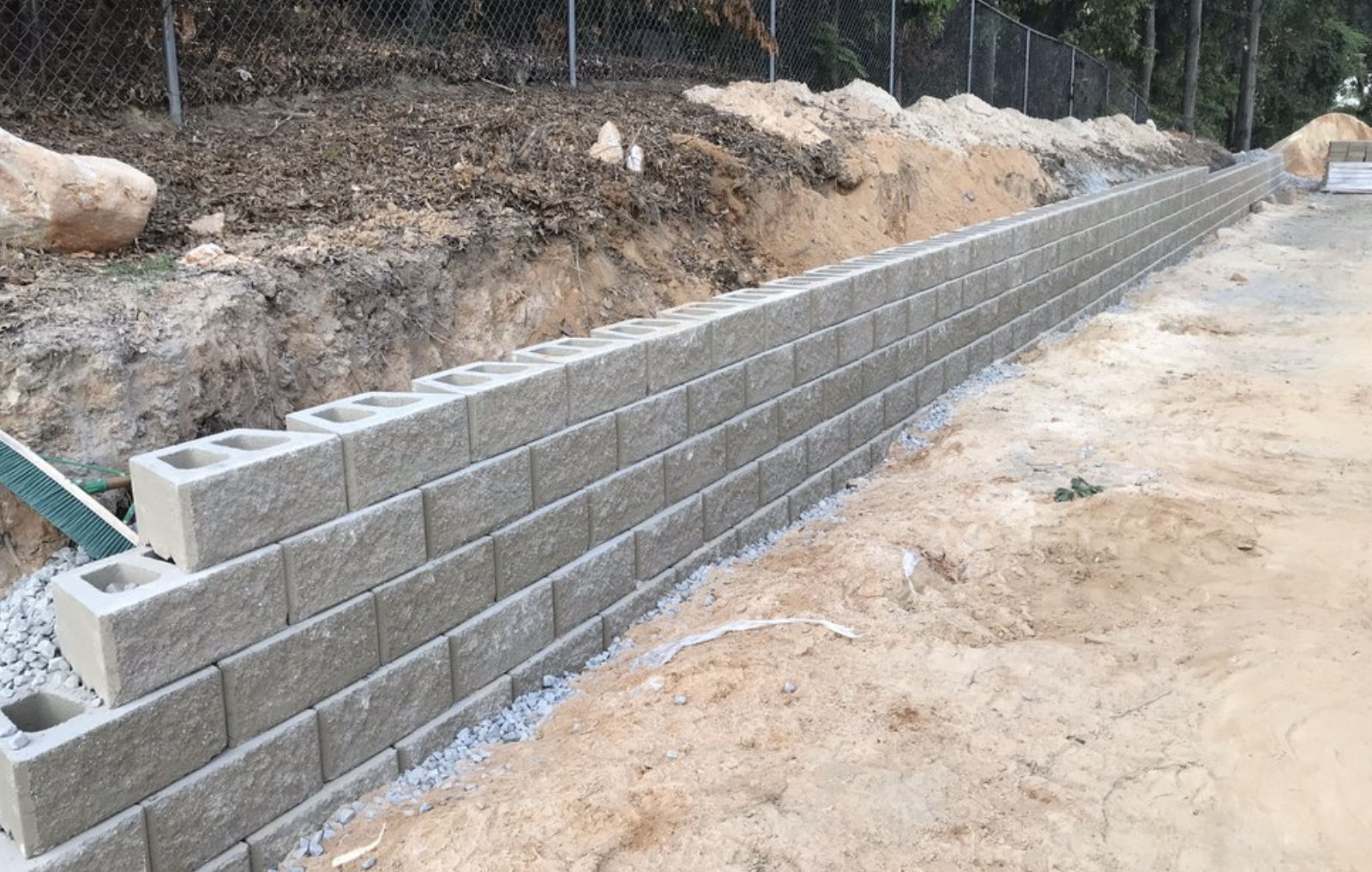 Construction of a concrete block retaining wall with some blocks at the beginning of the wall and others still to be installed, on a dirt surface with exposed soil behind.