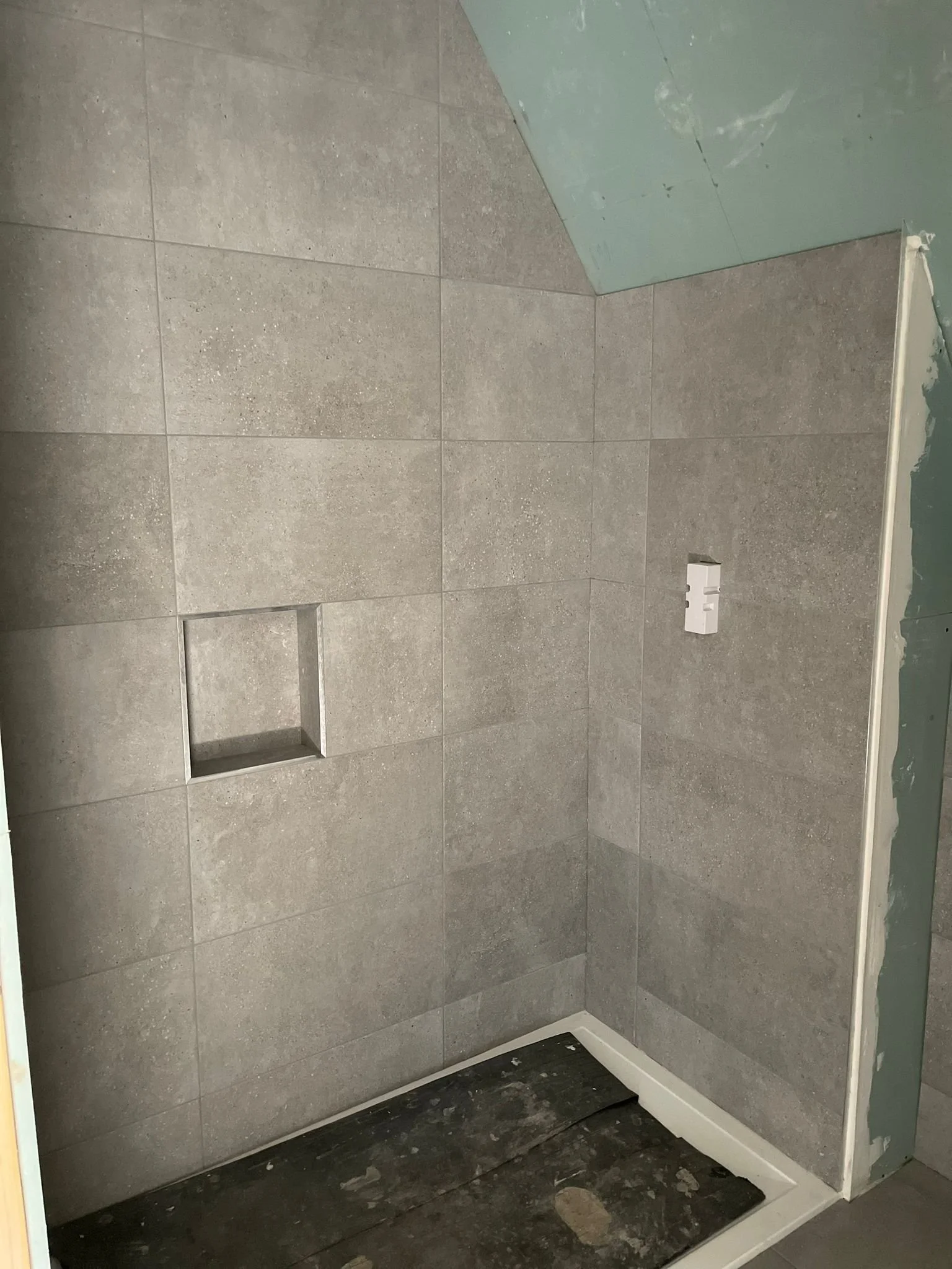 Bathroom shower area with gray tiles, a small built-in niche, and a partially installed wall with electrical outlet.