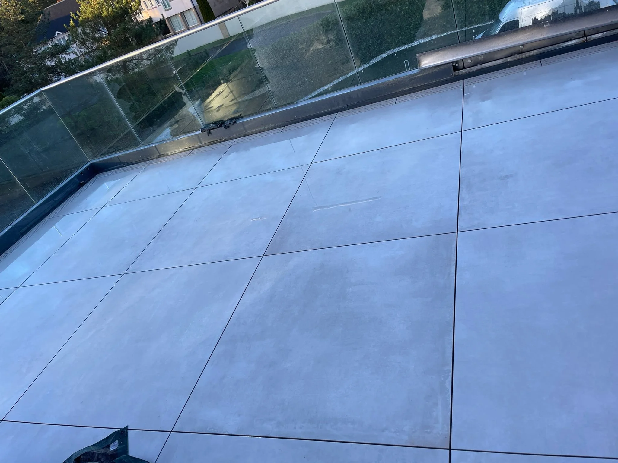 View of a balcony with large gray tiles, glass railing, and neighboring houses visible in the background.