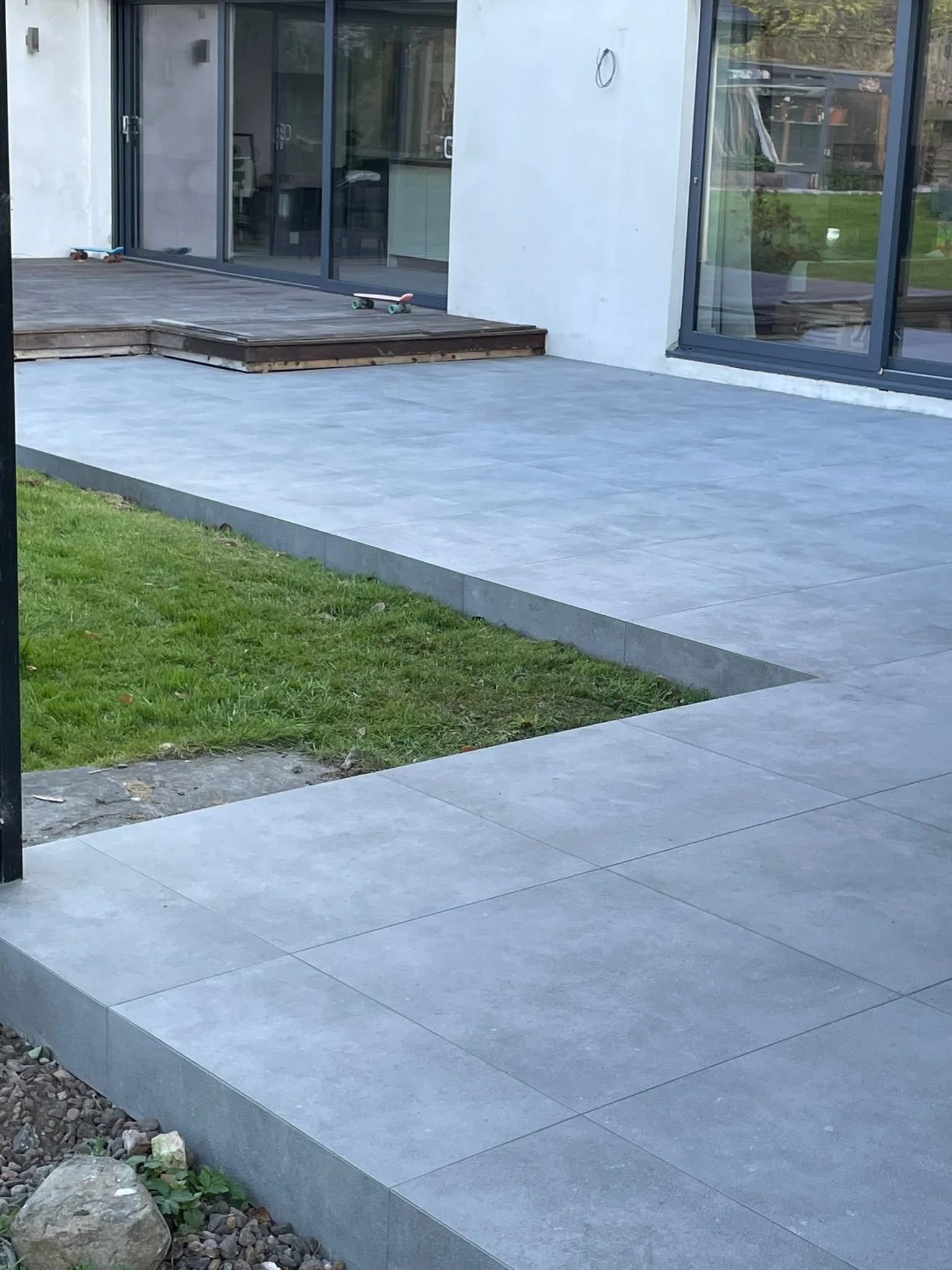 A newly installed grey tiled patio with a raised section leading to a sliding glass door. The patio is adjacent to a small grassy area and a rock garden.