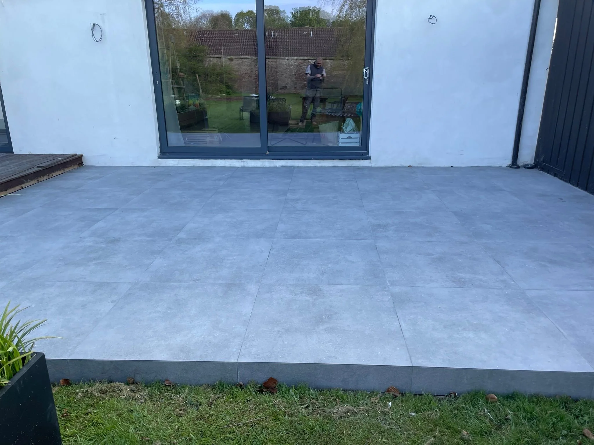 A newly installed grey tiled patio adjacent to a house with glass sliding doors, with a small wooden deck on the left side and a grassy area in the foreground, and the reflection of a person taking the photo visible in the glass.
