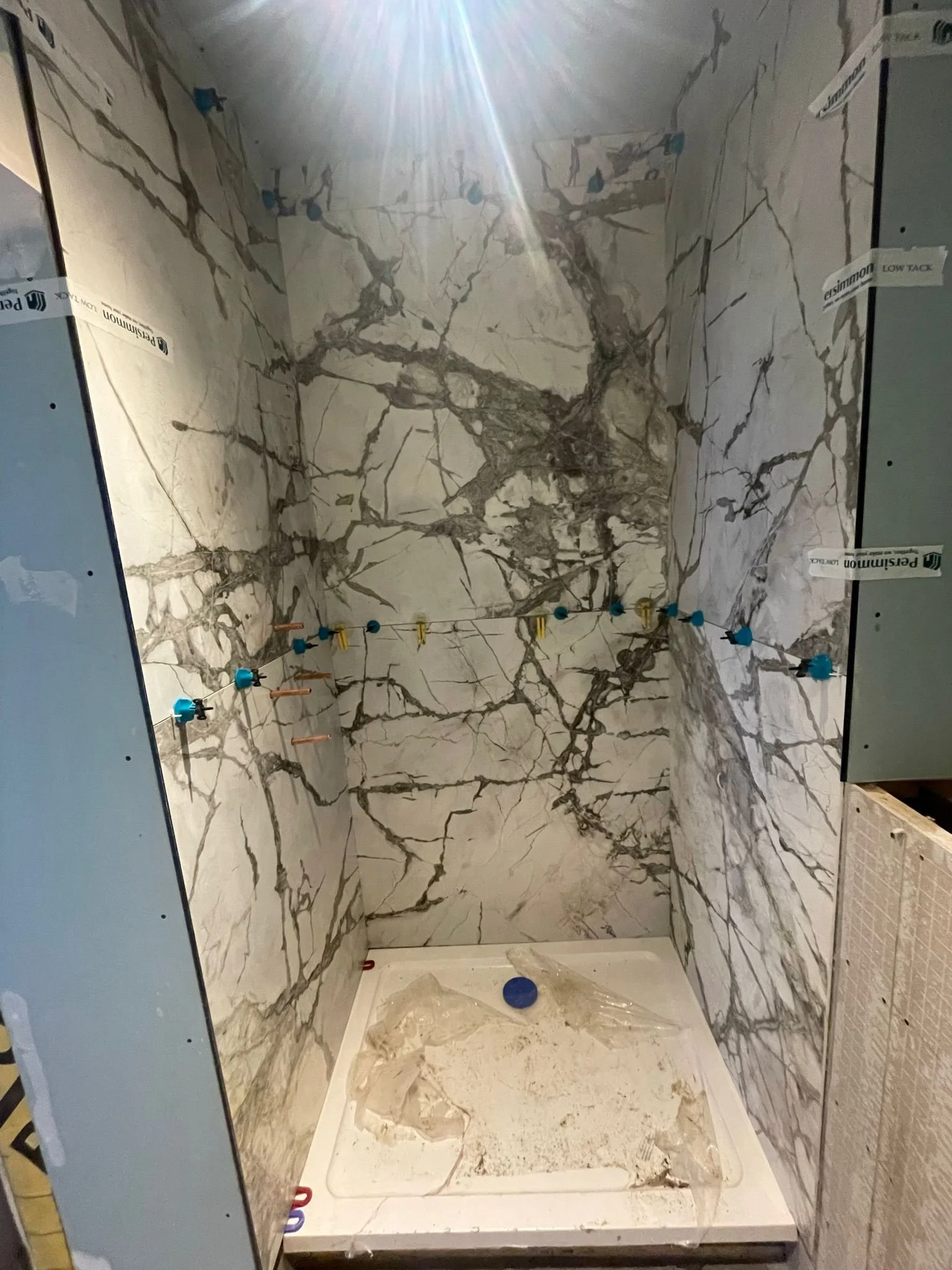 Bathroom shower area under construction with marble wall tiles and an unfinished shower base with plastic covering. Pipes and fixtures are not yet installed.