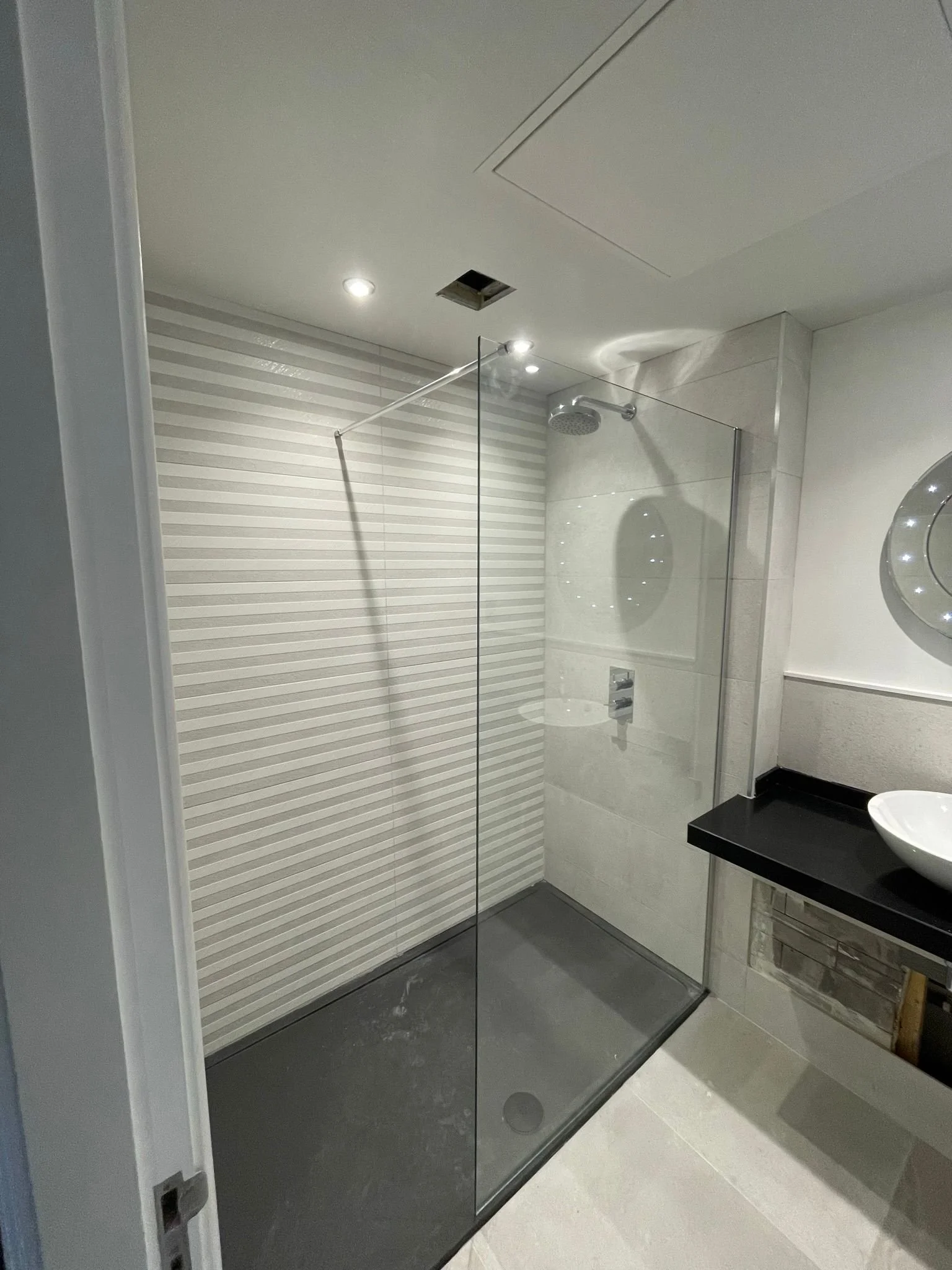 Modern bathroom with a walk-in shower featuring a glass partition, a rainfall showerhead, light-colored tiled walls, a black countertop with a white vessel sink, and a circular mirror with built-in lighting.