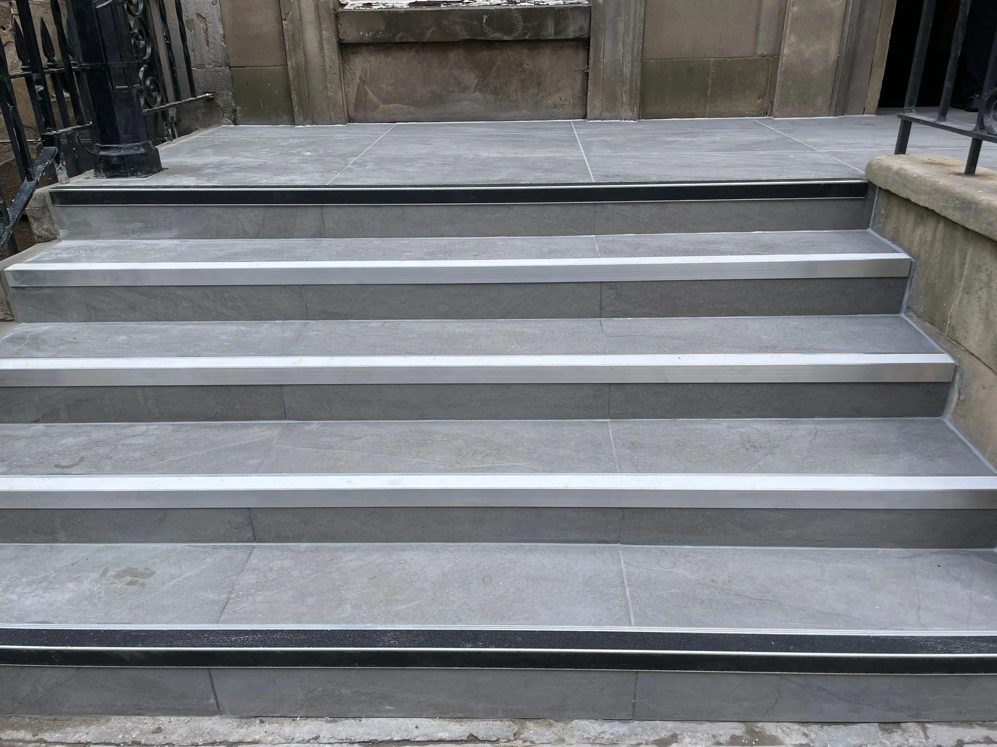 A set of outdoor grey tiled stairs with metal safety strips along each step, leading to a wooden door with a stone wall on the right side and black metal railings on the left side.