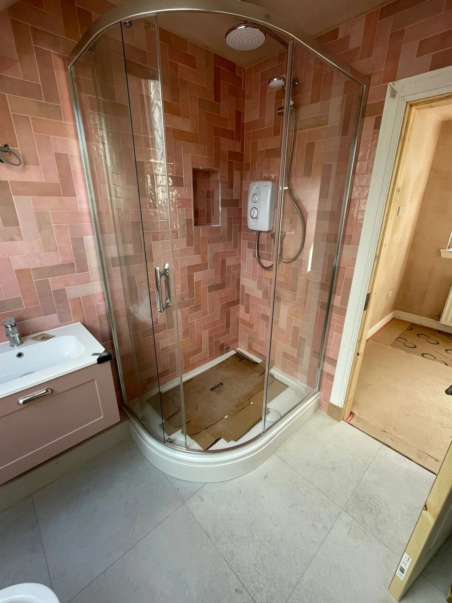 A corner shower with pink brick-patterned tiles, a glass door, and a shower panel with controls and a handheld showerhead. There is a built-in niche in the wall and a floor drain, with an area of cardboard on the shower base.