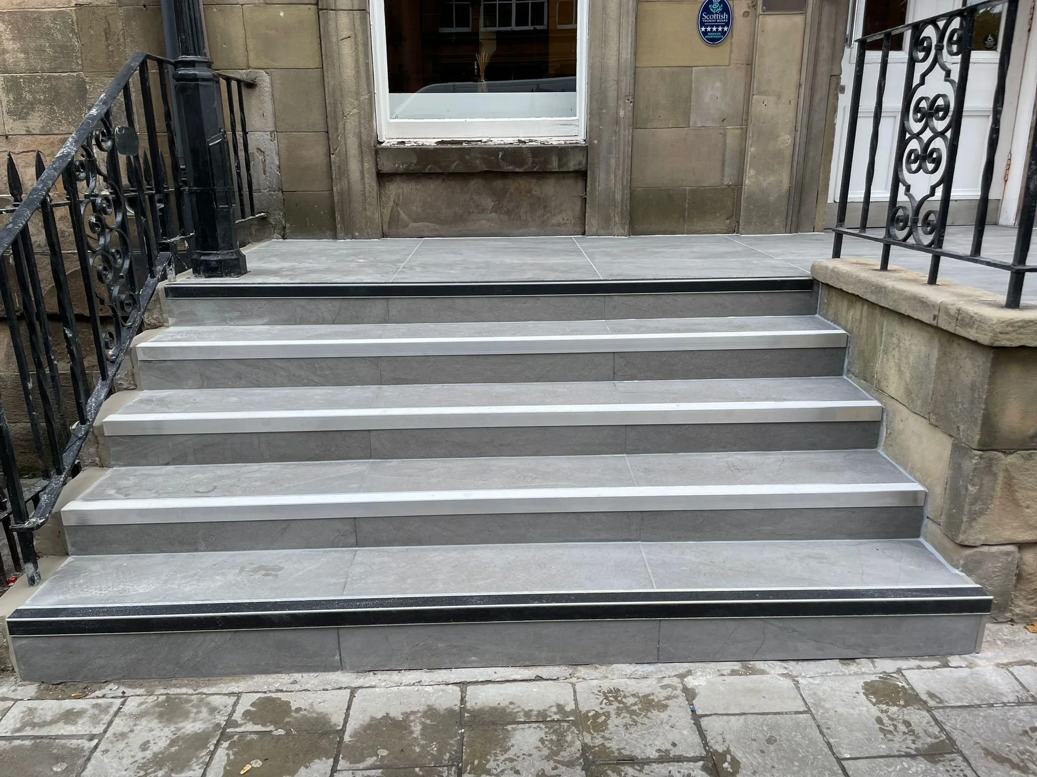 Five concrete steps with metal edges and black and beige striped lines lead up to a building entrance with a glass door and a large window, flanked by black metal railings on both sides.