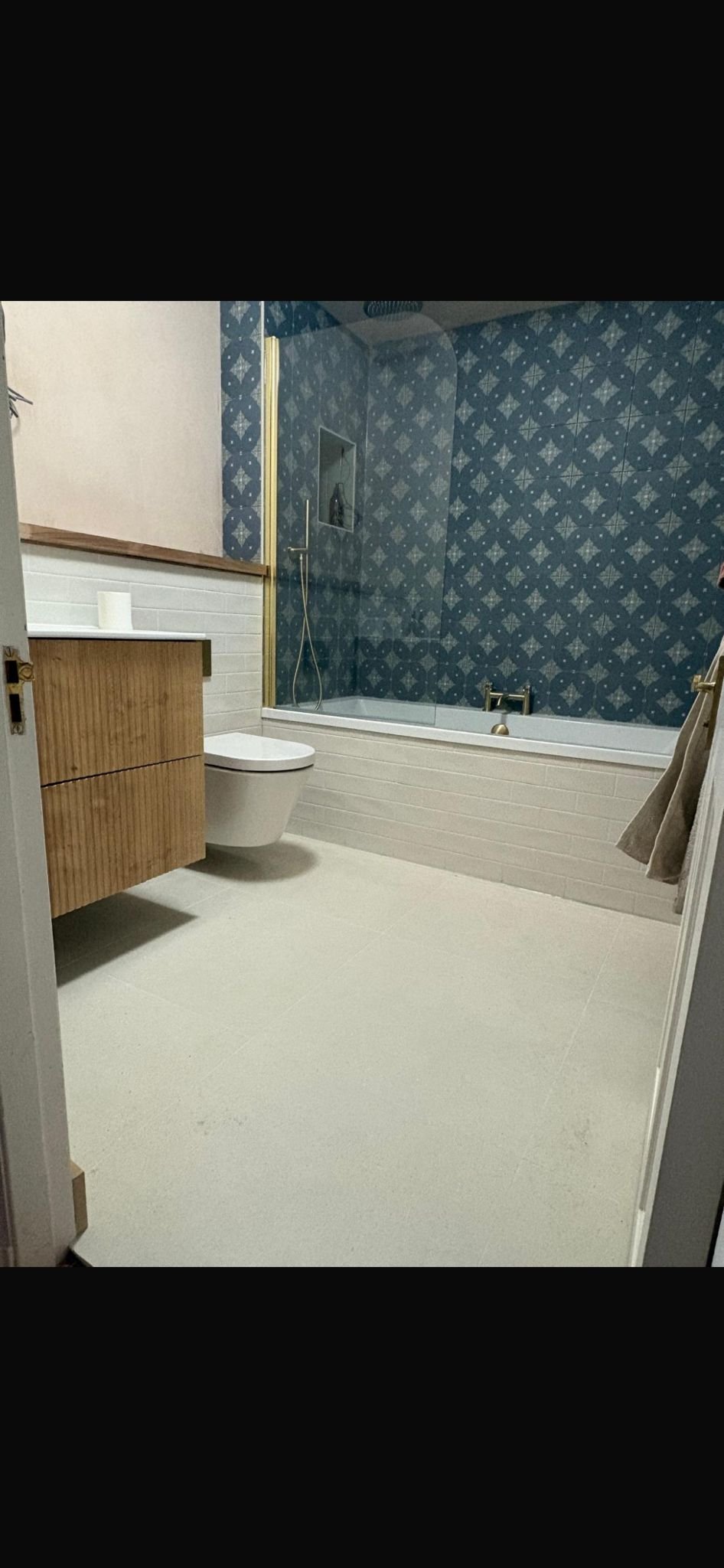 Bathroom with a wooden vanity, wall-mounted toilet, blue patterned tiled wall in the shower area, bathtub, towel hanging on the side, and a glass shower screen.