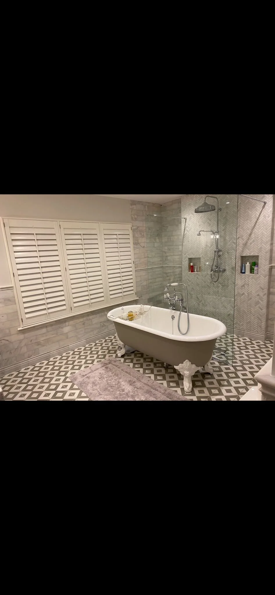 Elegant bathroom with vintage-style clawfoot bathtub, patterned floor tiles, glass shower enclosure with built-in shelves, and a window with white shutters.