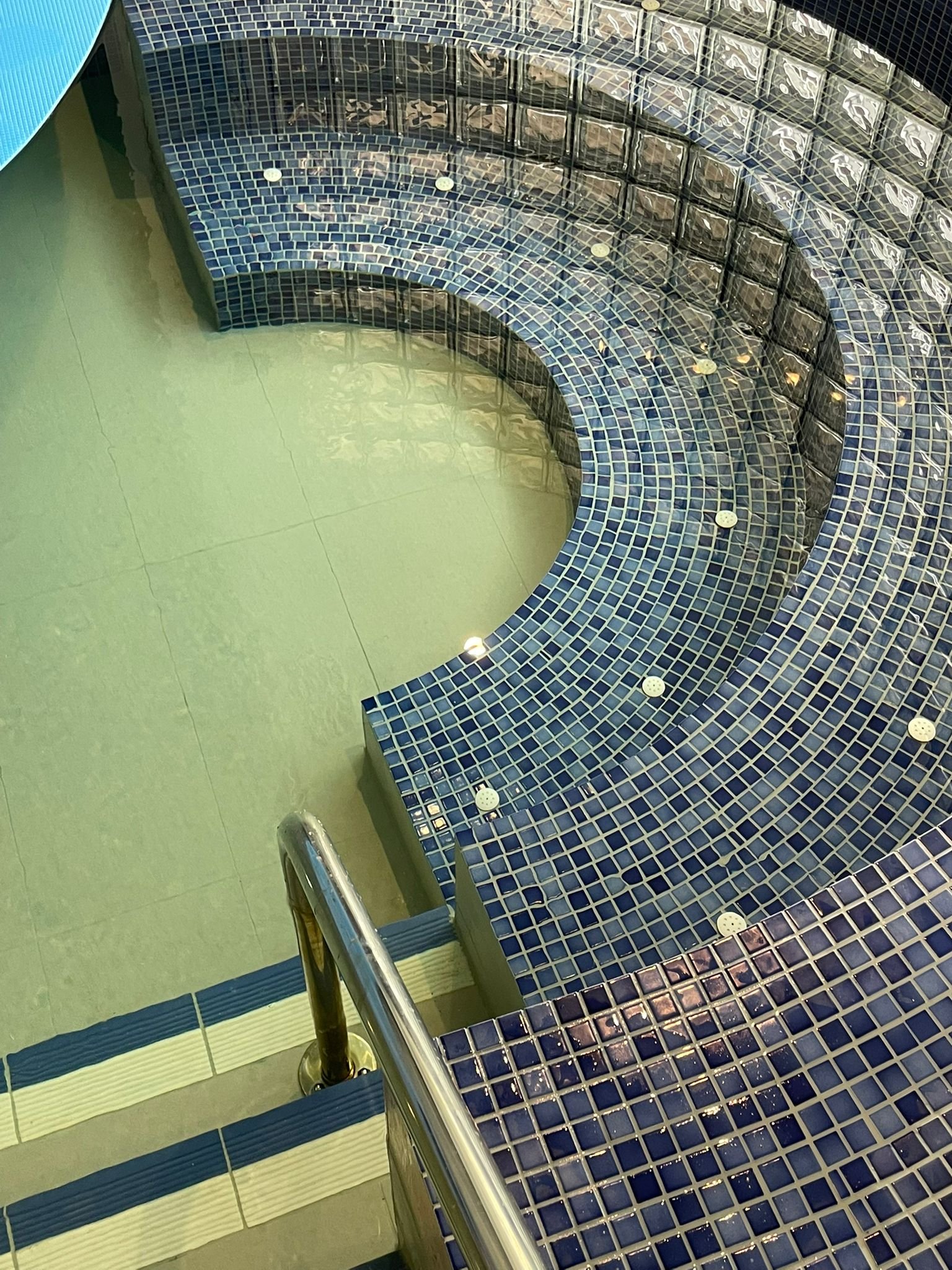 Indoor hot tub with blue mosaic tiles, a stainless steel handrail, and steps leading into the water.