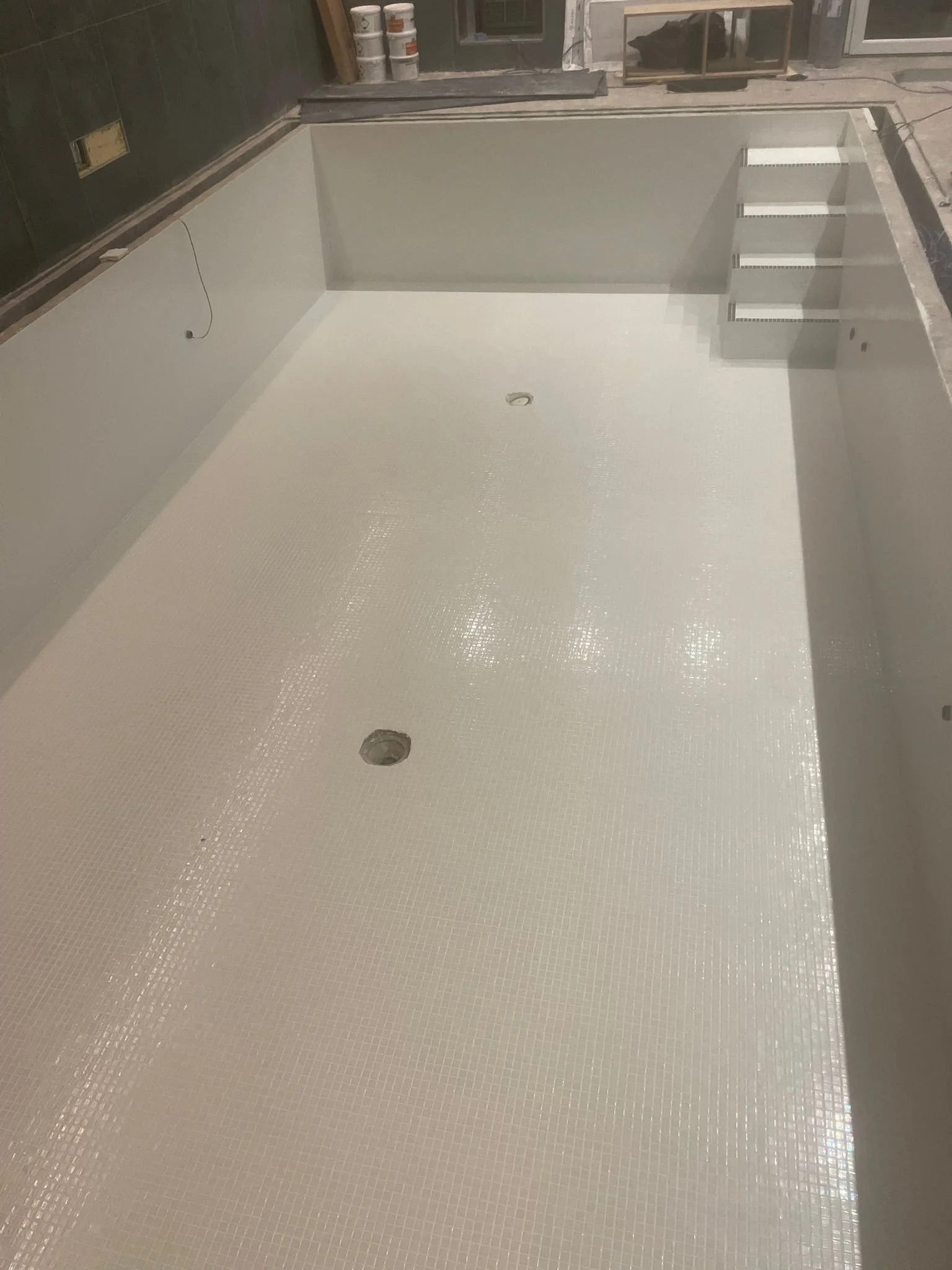 Empty indoor pool or hot tub with a white tiled floor, two drain openings, unfinished walls, and some construction supplies in the background.