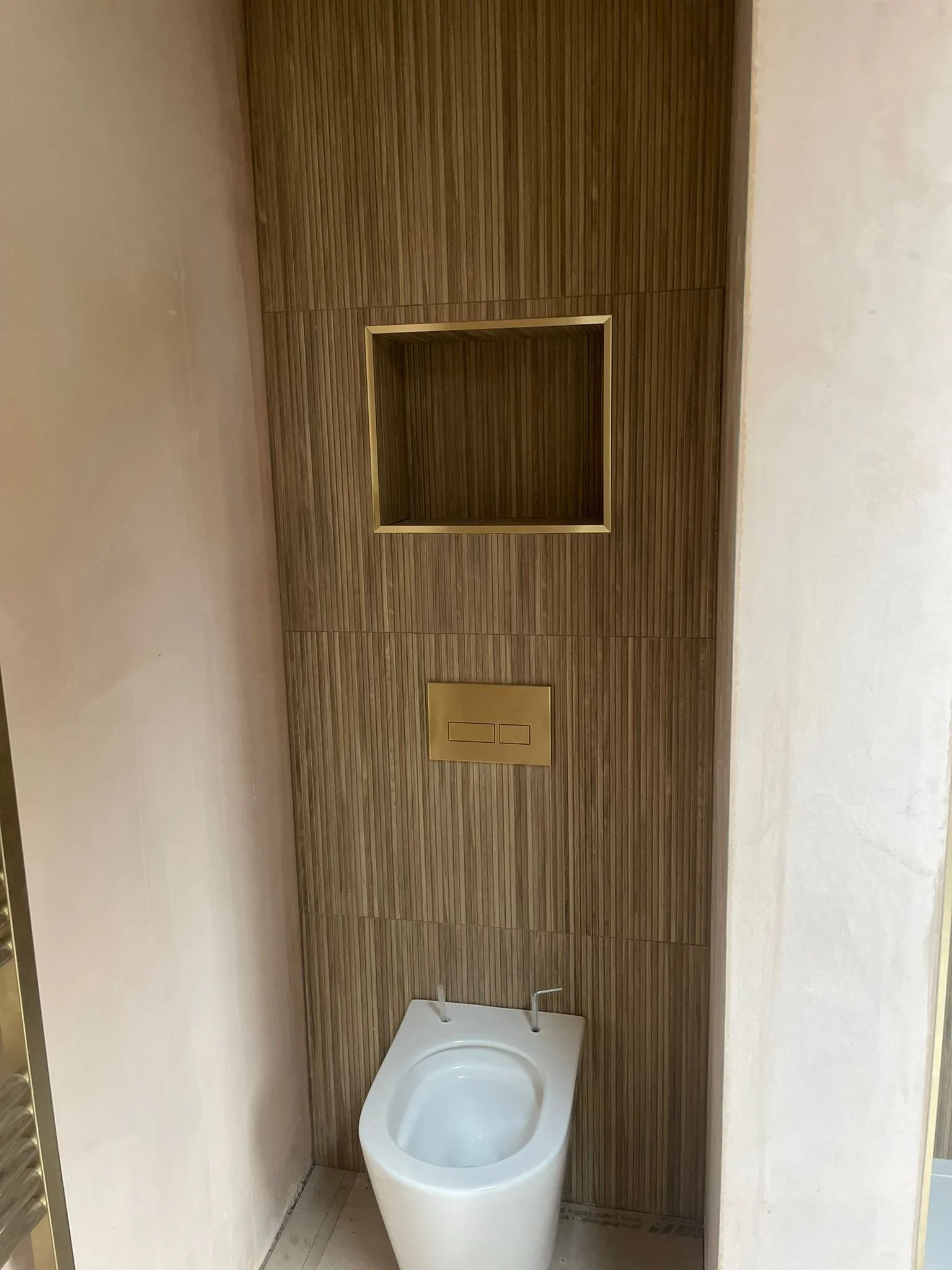 Modern bathroom with wooden wall paneling, a built-in shelf with gold trim, and a white bidet below a gold flush plate.