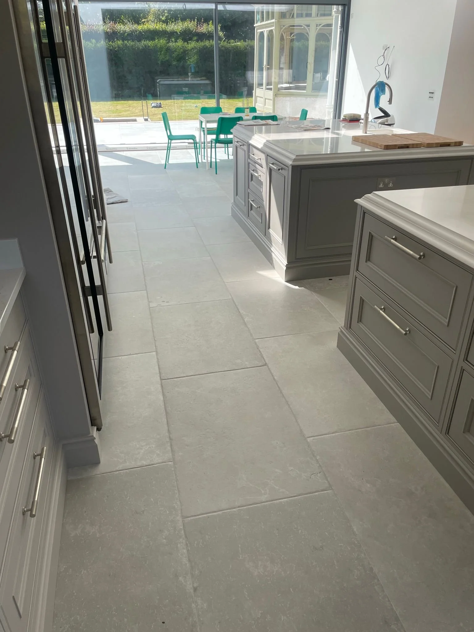 View of a modern kitchen with gray cabinets, large tiled floor, and a sliding glass door leading to an outdoor area with a patio and green lawn, with teal dining chairs outside.