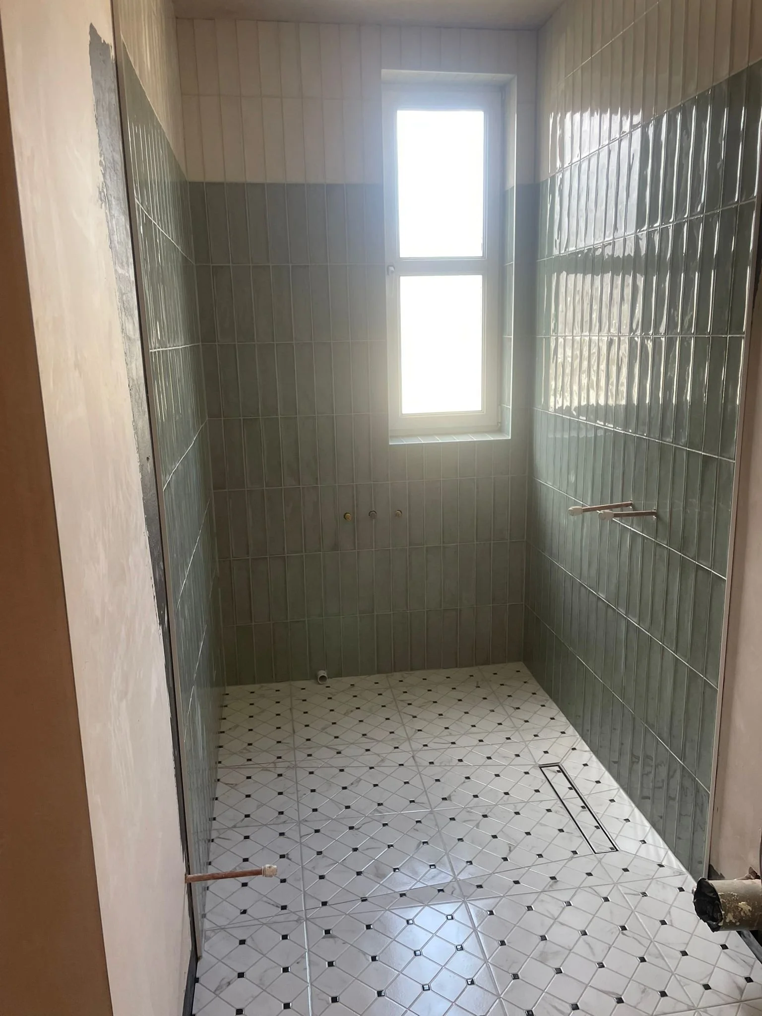 Empty bathroom with green tiled walls, white patterned floor tiles, a window, and plumbing fixtures for a shower or bathtub.