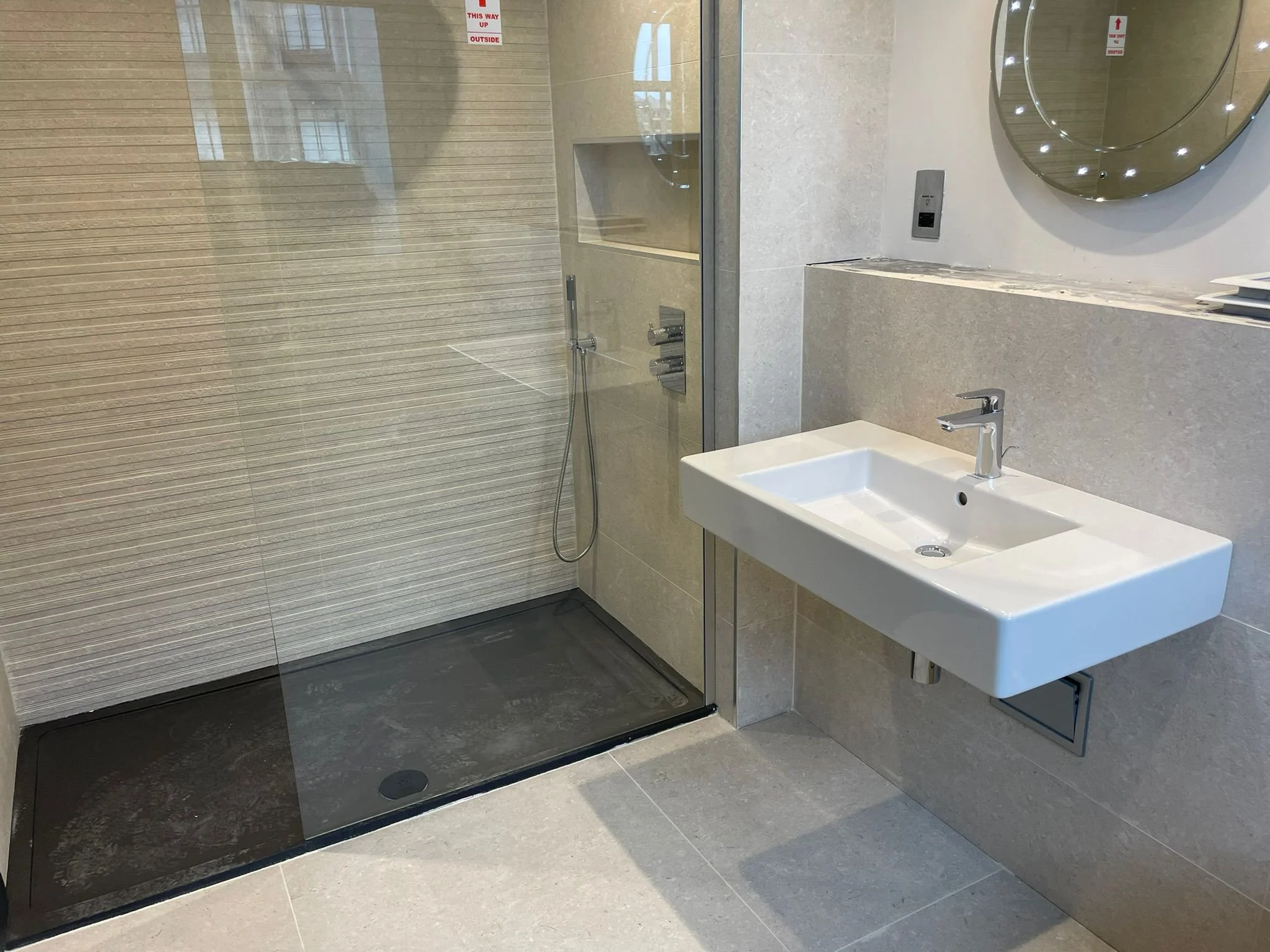 A modern bathroom with a walk-in shower featuring beige tiled walls and a black shower base. To the right, a white rectangular wall-mounted sink with a chrome faucet is installed on a beige tiled wall, above a small access panel. A large round mirror