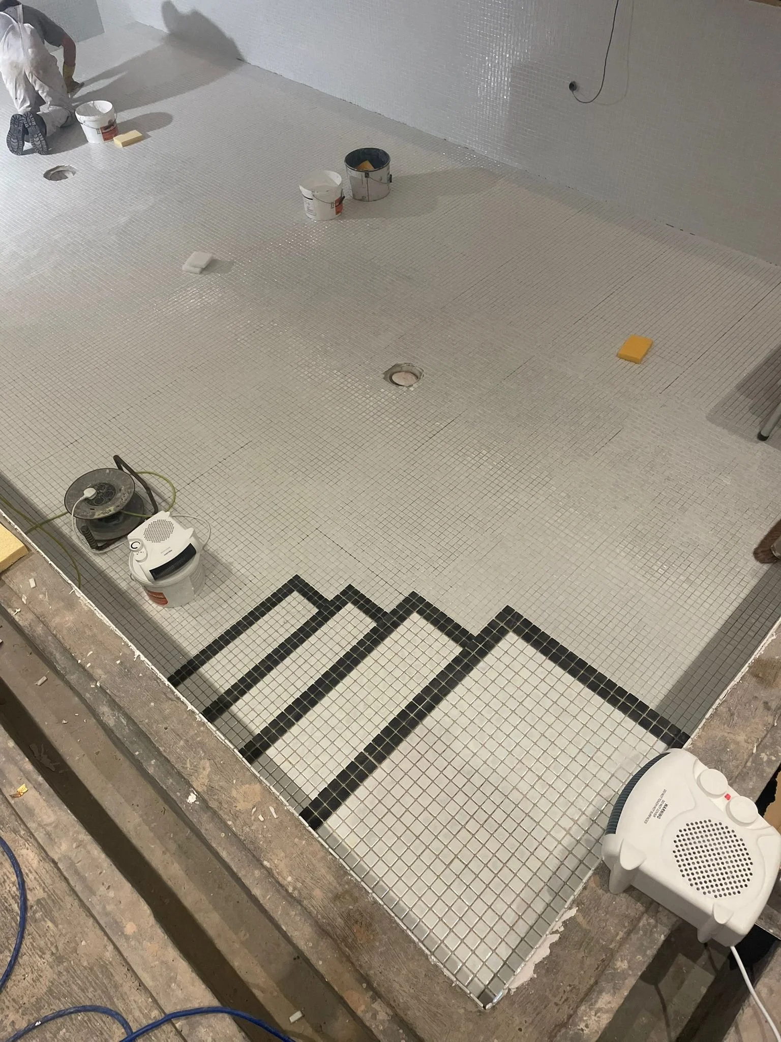 Under construction of a tiled floor with black border tiles forming a geometric pattern, construction materials, and tools scattered around.