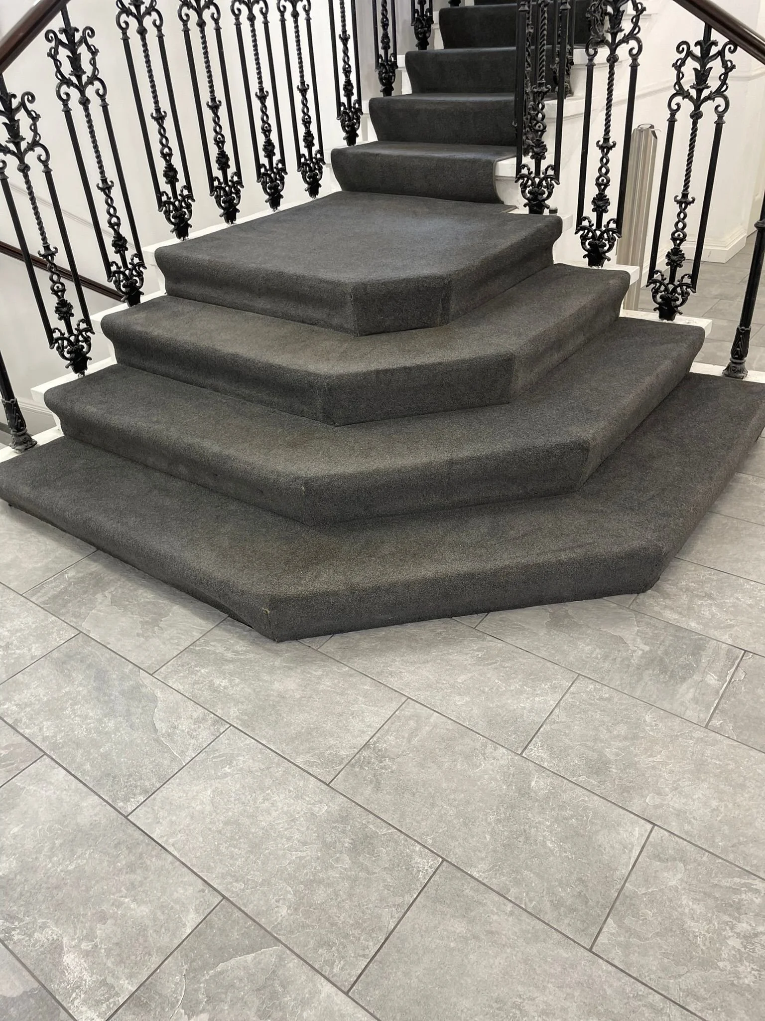 Interior of a house featuring a staircase with black wrought iron railing, gray carpeted stairs, and gray floor tiles.