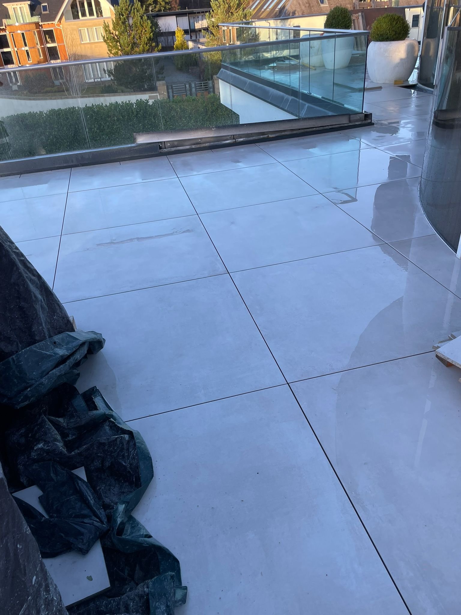 A modern balcony with a glass railing, white tiled floor, potted plants, and a view of neighboring buildings with trees in the background.