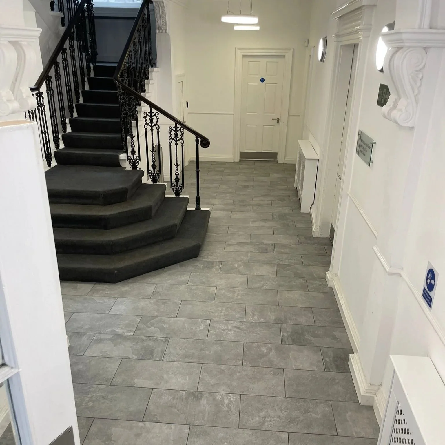 Interior of a building with gray tiled floor, white walls, a staircase with black railing, and a closed white door in the background.