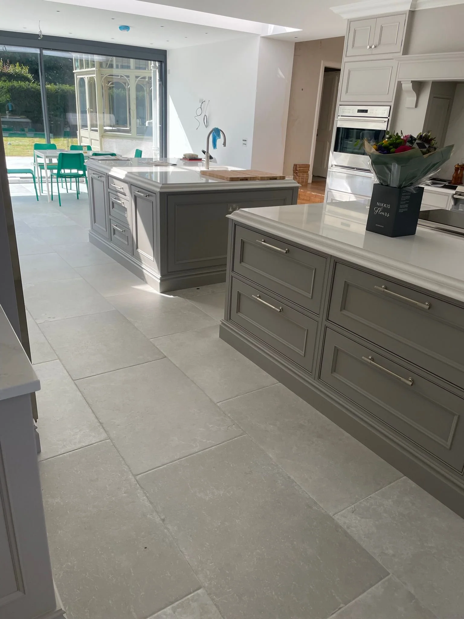 Modern kitchen with gray cabinets, white countertops, and large light-colored floor tiles. A sliding glass door leads to an outdoor area with a dining table and chairs. There are flowers on the counter and built-in appliances in the background.