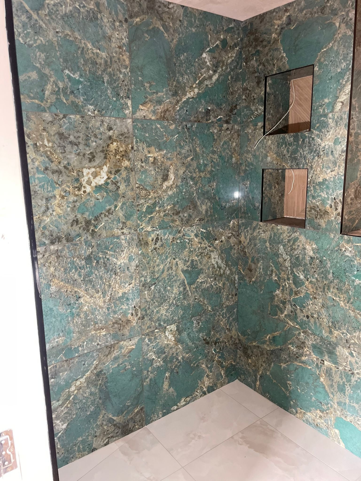 A corner of a bathroom with large green and brown marble tiles, two built-in wall shelves with wooden backing, and a white tiled floor.