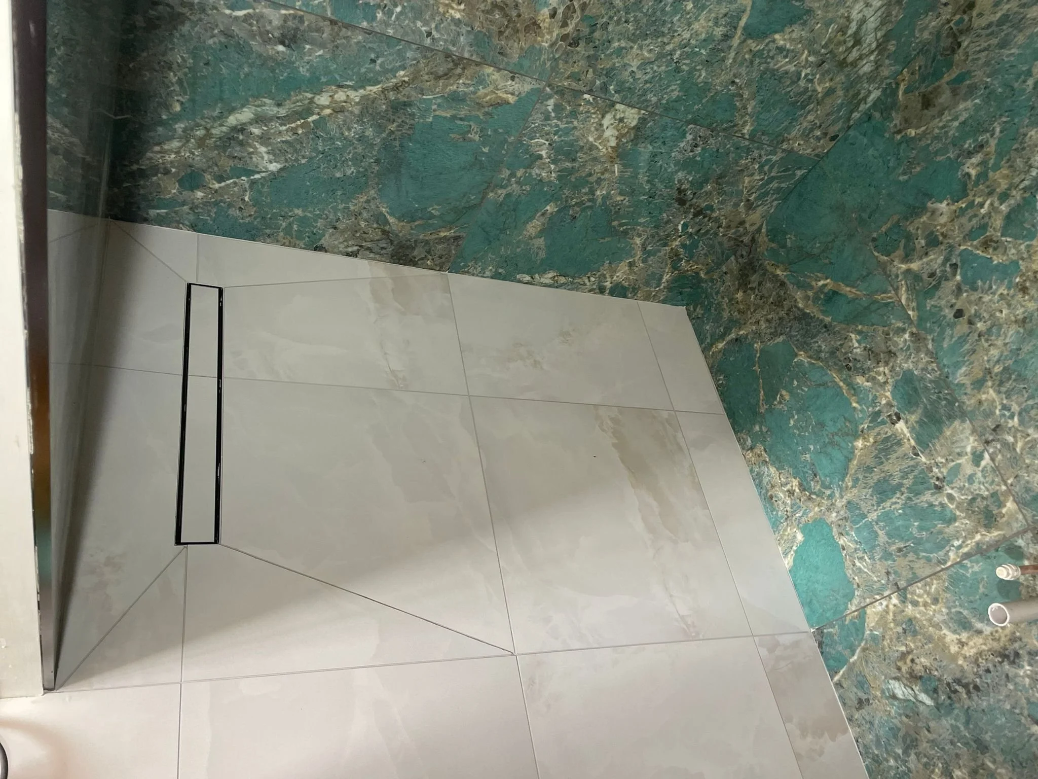 Close-up of a corner in a bathroom showcasing different tiled floors: white marble tiles on the lower part of the wall and floor, and green marbled tiles on the upper wall and surrounding areas.