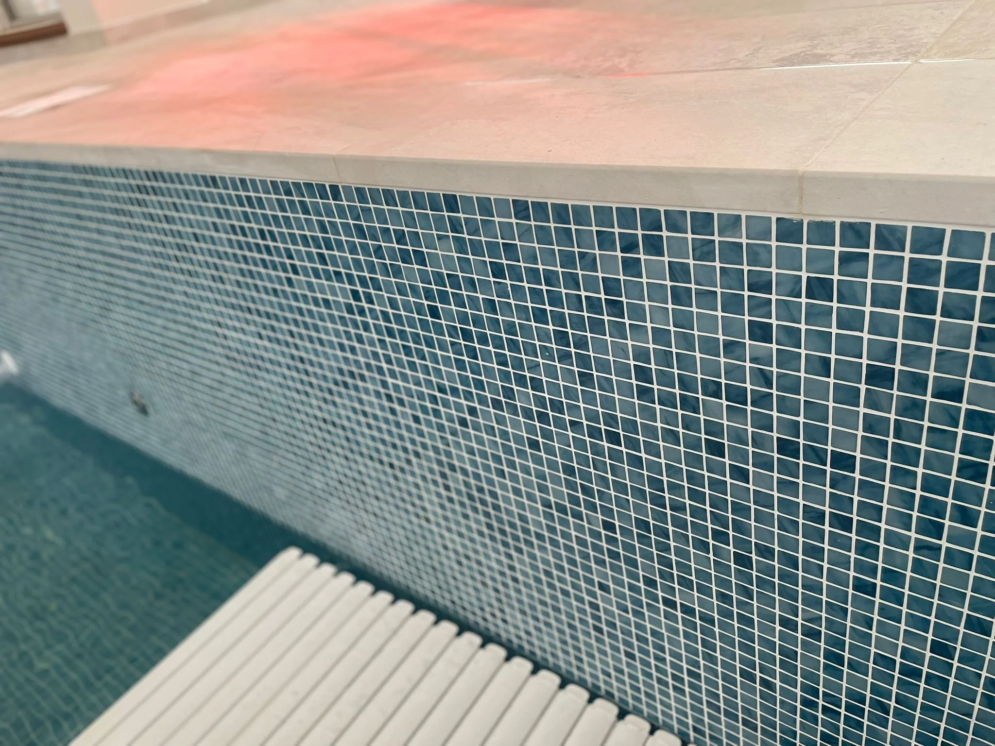 Close-up of a tiled pool wall with small dark blue mosaic tiles and a metal drain cover at the bottom.