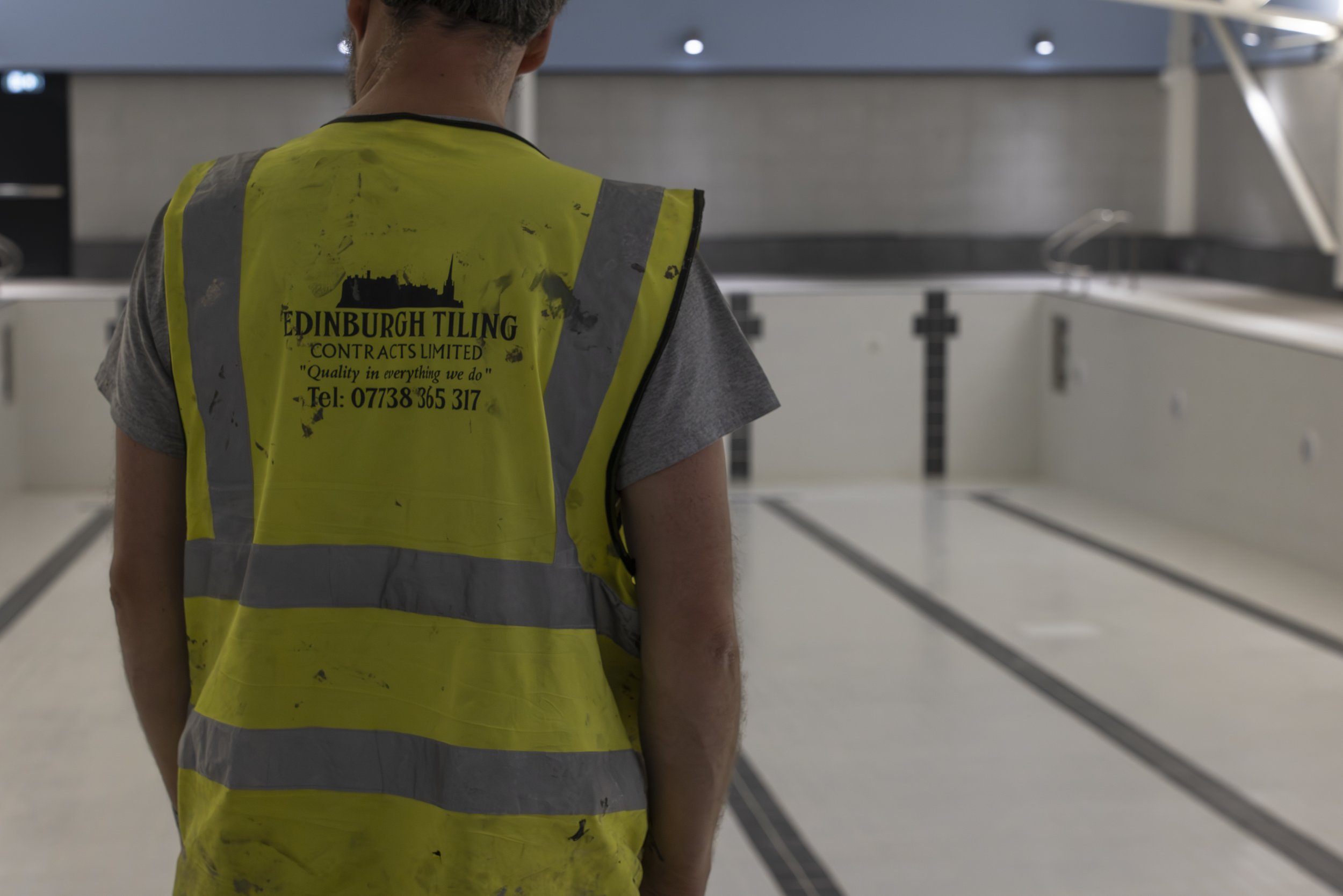 A construction worker wearing a yellow safety vest with black text, standing in an empty room that appears to be under renovation, with white walls and black striping on the floor.