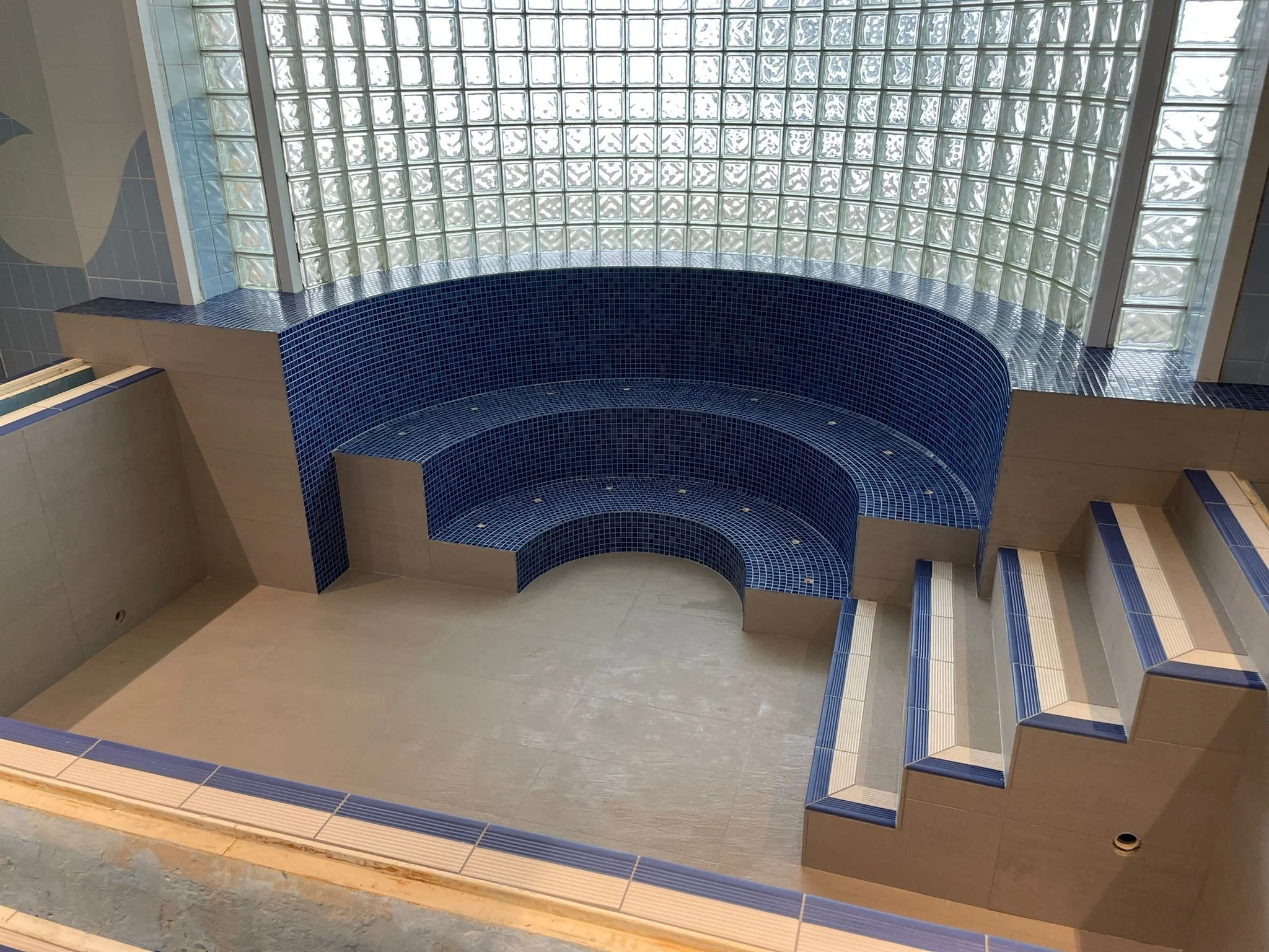 Interior with a curved blue tiled seating area with three steps leading up to it, surrounded by large glass block windows.