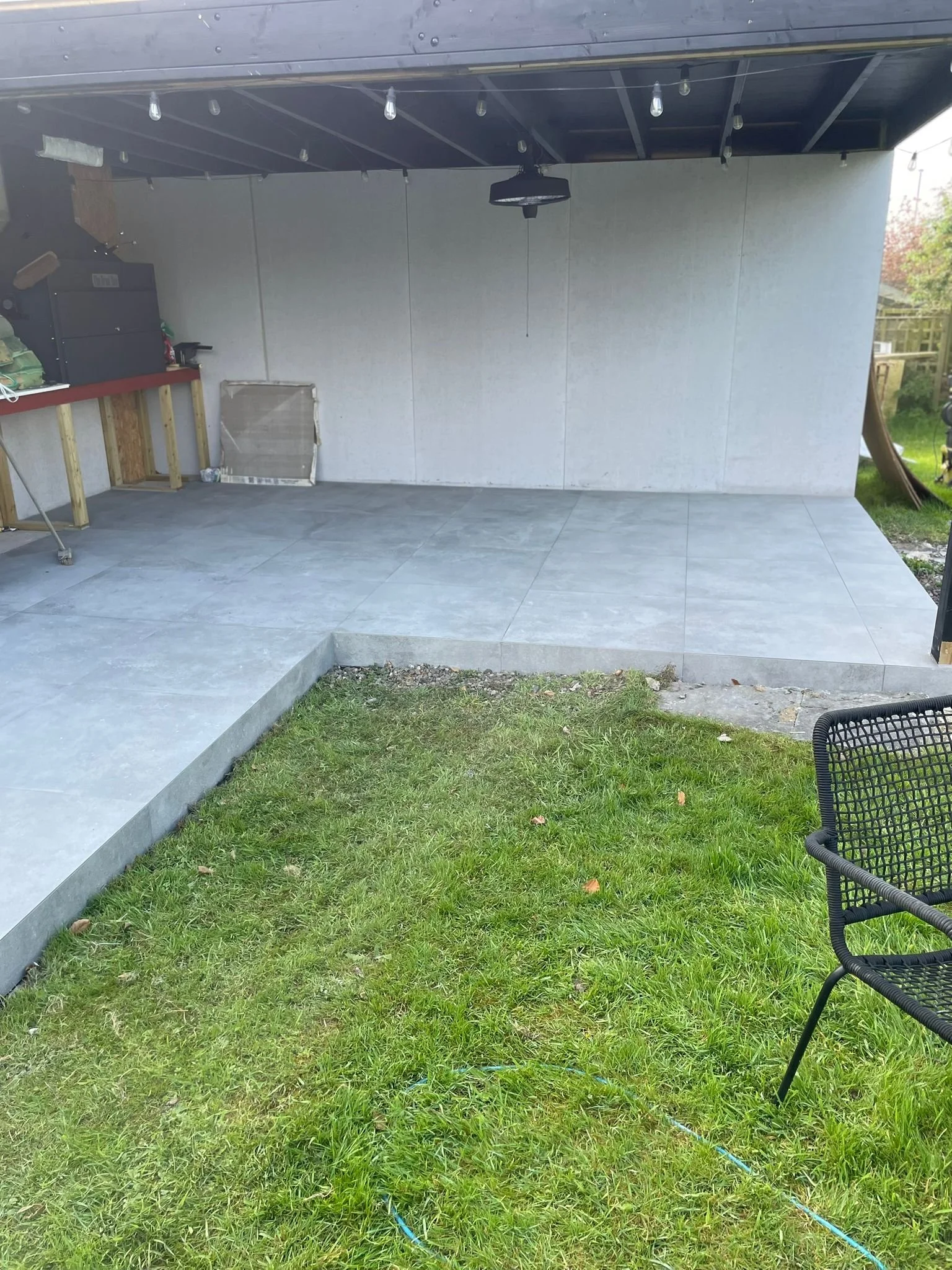 Empty backyard patio with grey tile flooring, a black outdoor chair, and a grass lawn with some weeds and brown leaves.