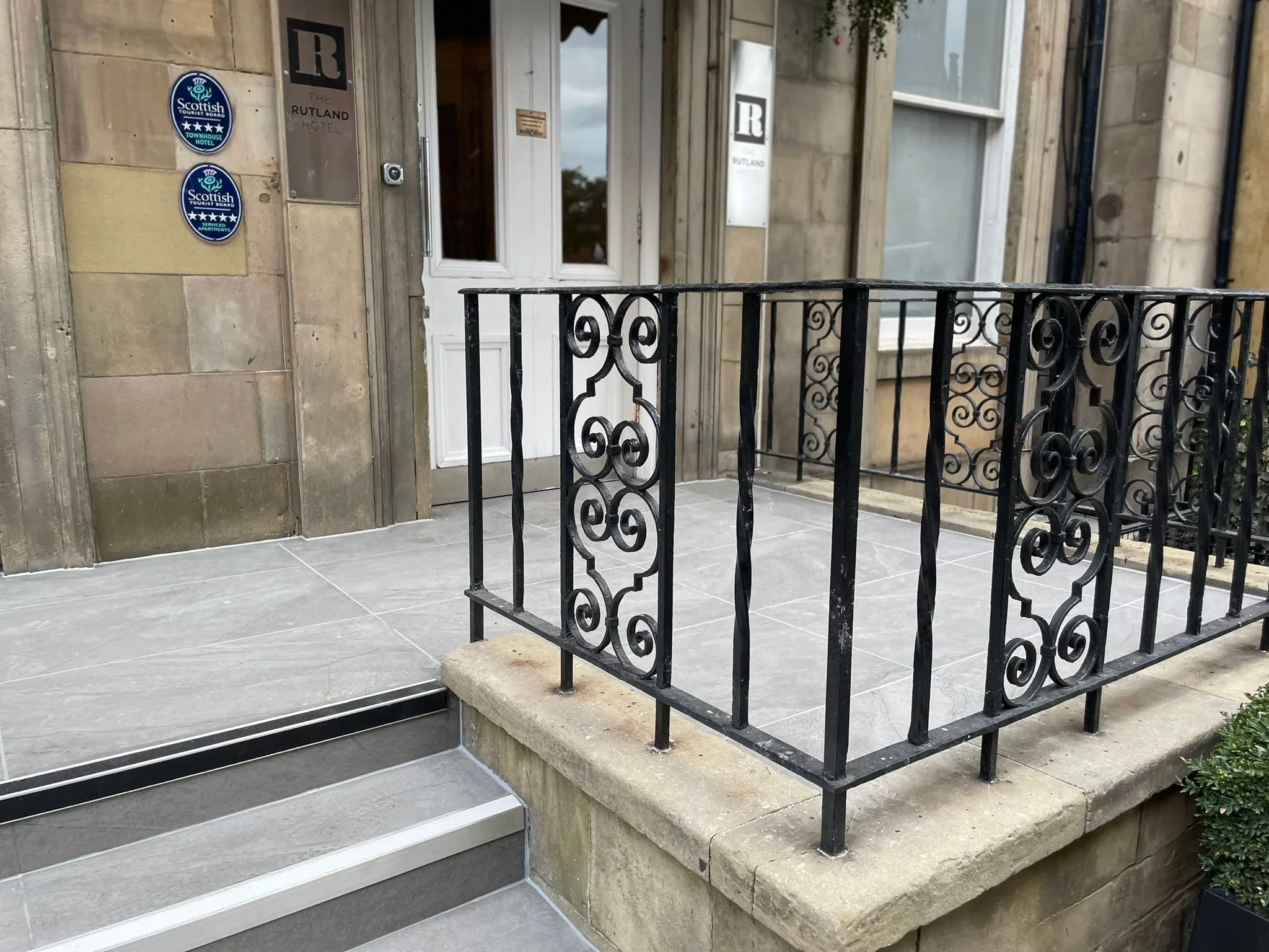 Small front porch with ornate black iron railing, gray tiles, and steps leading to white front door of building.