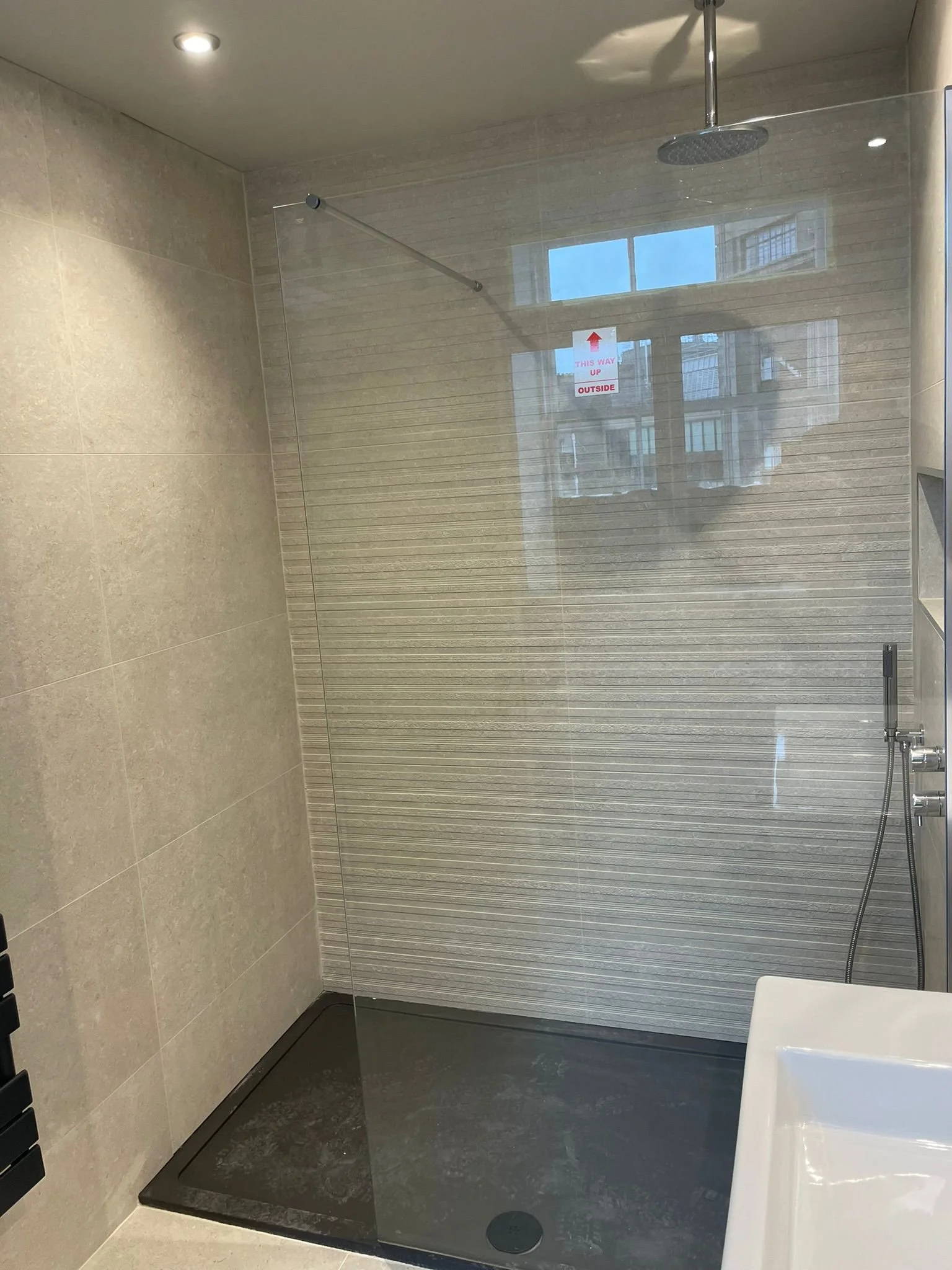 Modern shower area with a large rainfall showerhead, a glass partition, beige tiled walls, and a black textured shower base, with a window reflecting buildings outside standing behind a window sticker.
