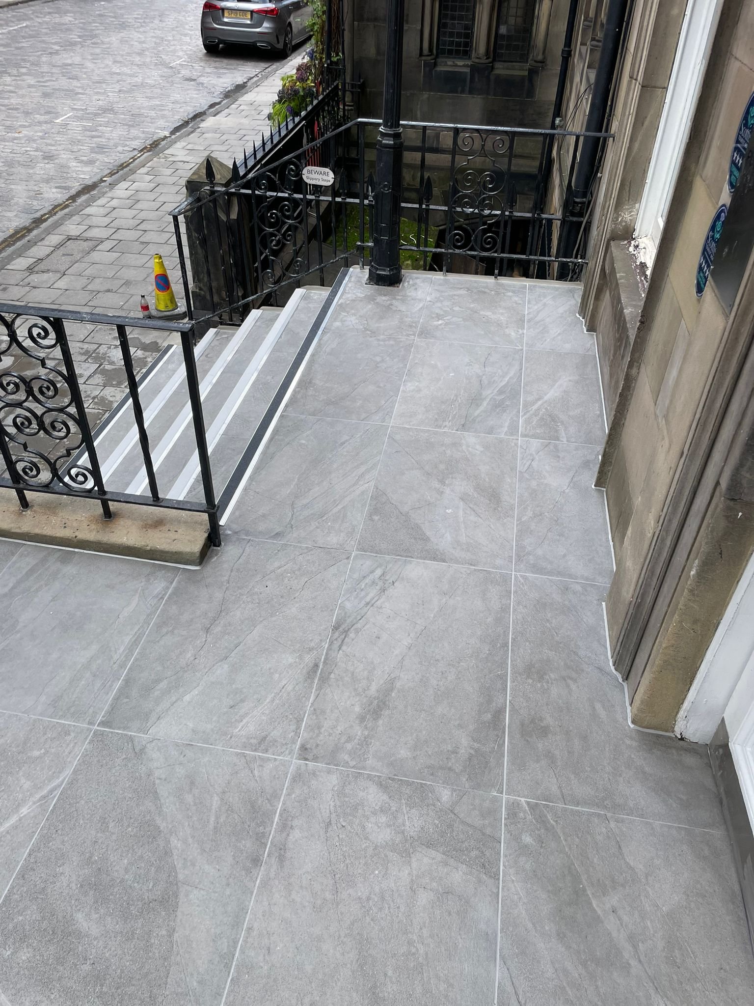 Outdoor building entrance with gray tiled floor, black wrought iron handrails, staircase leading down, and a small yellow cone on the sidewalk.