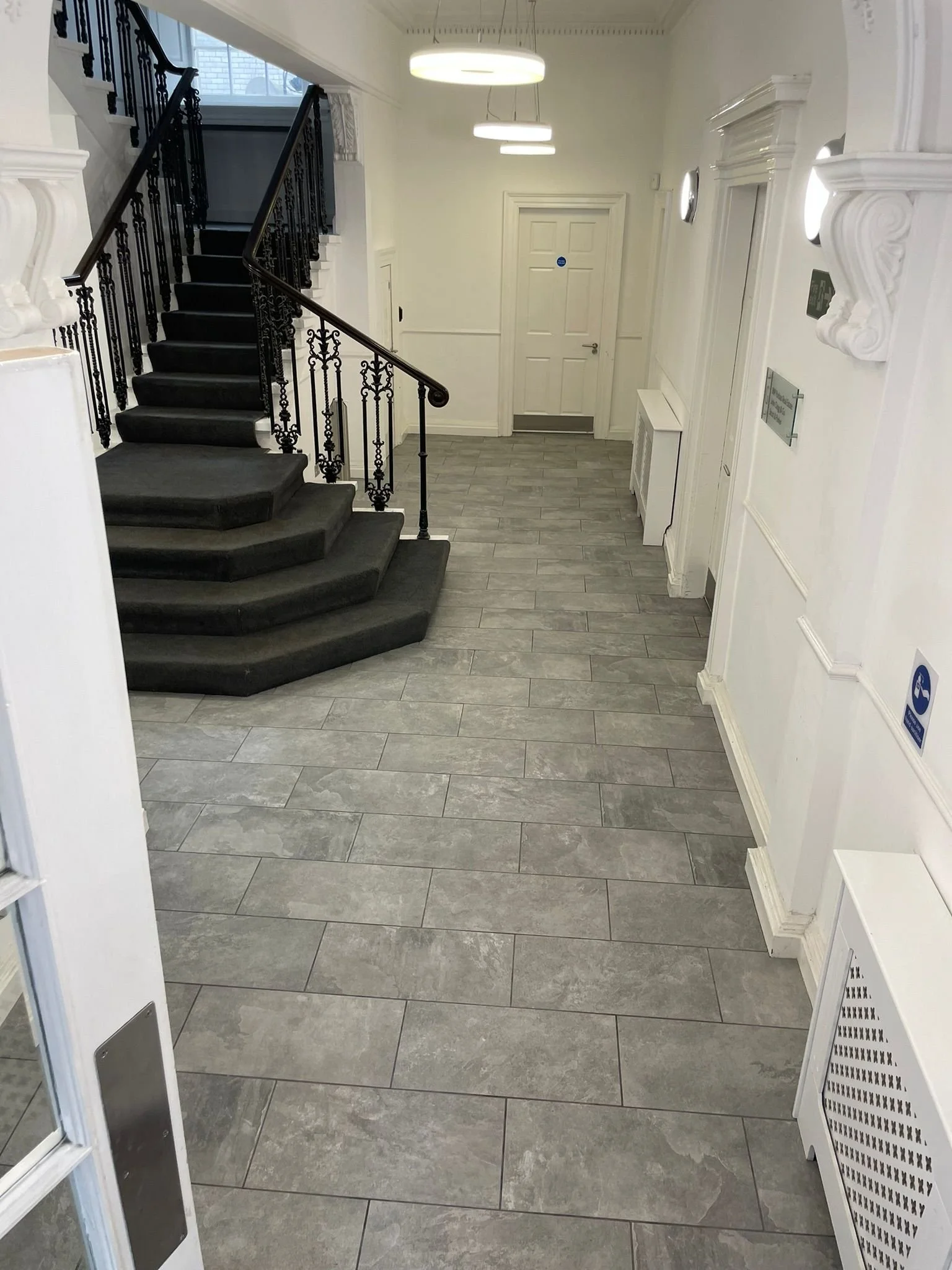 Interior view of a building lobby with gray tiled flooring, white walls, decorative moldings, a staircase with black wrought iron railing and carpeted steps, and a white door against the far wall.