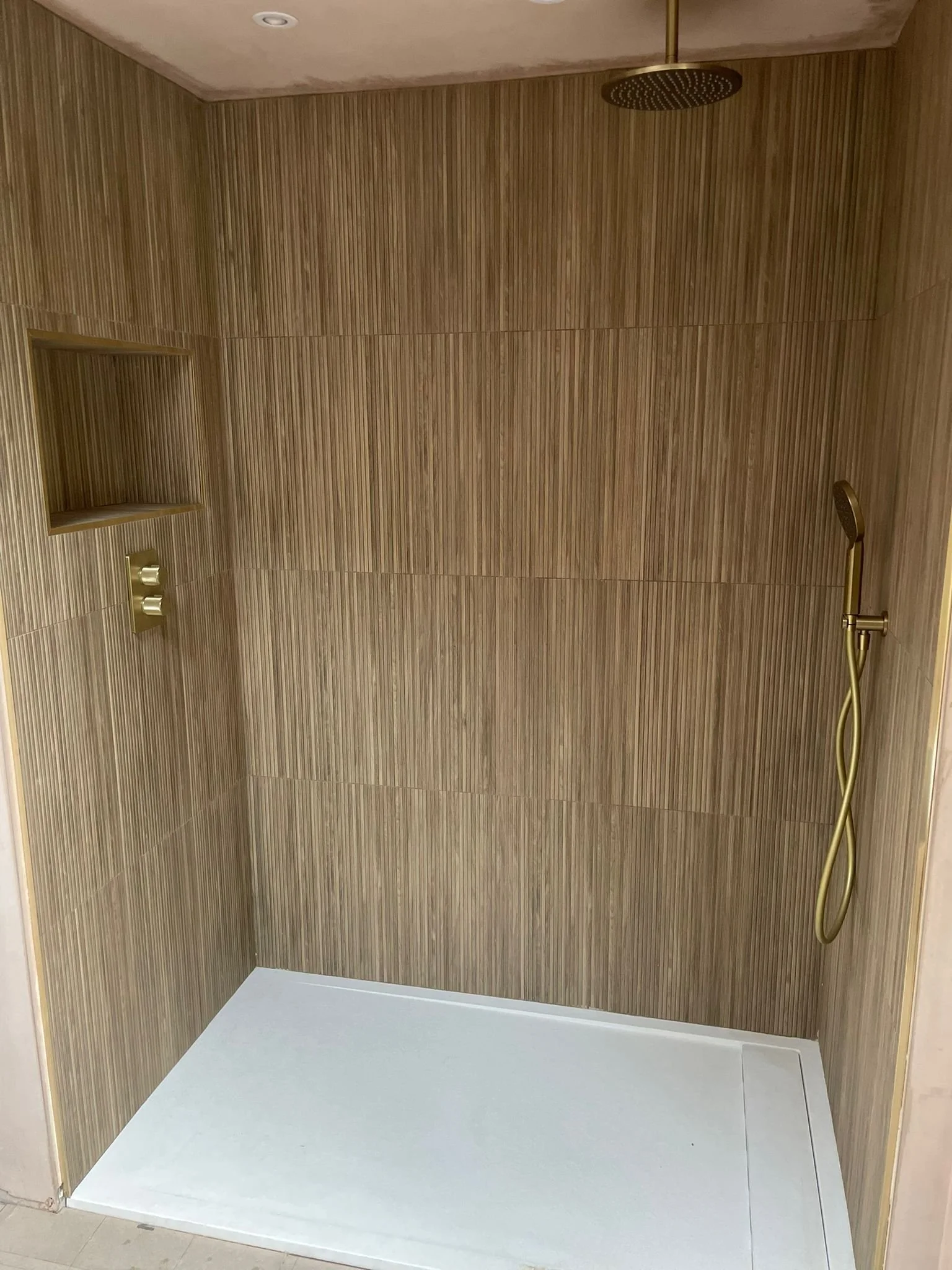 Empty walk-in shower with brown wood-textured tile walls, a black rainfall showerhead mounted on the ceiling, a golden handheld showerhead on a flexible hose, a built-in wall niche, and a white shower floor base.