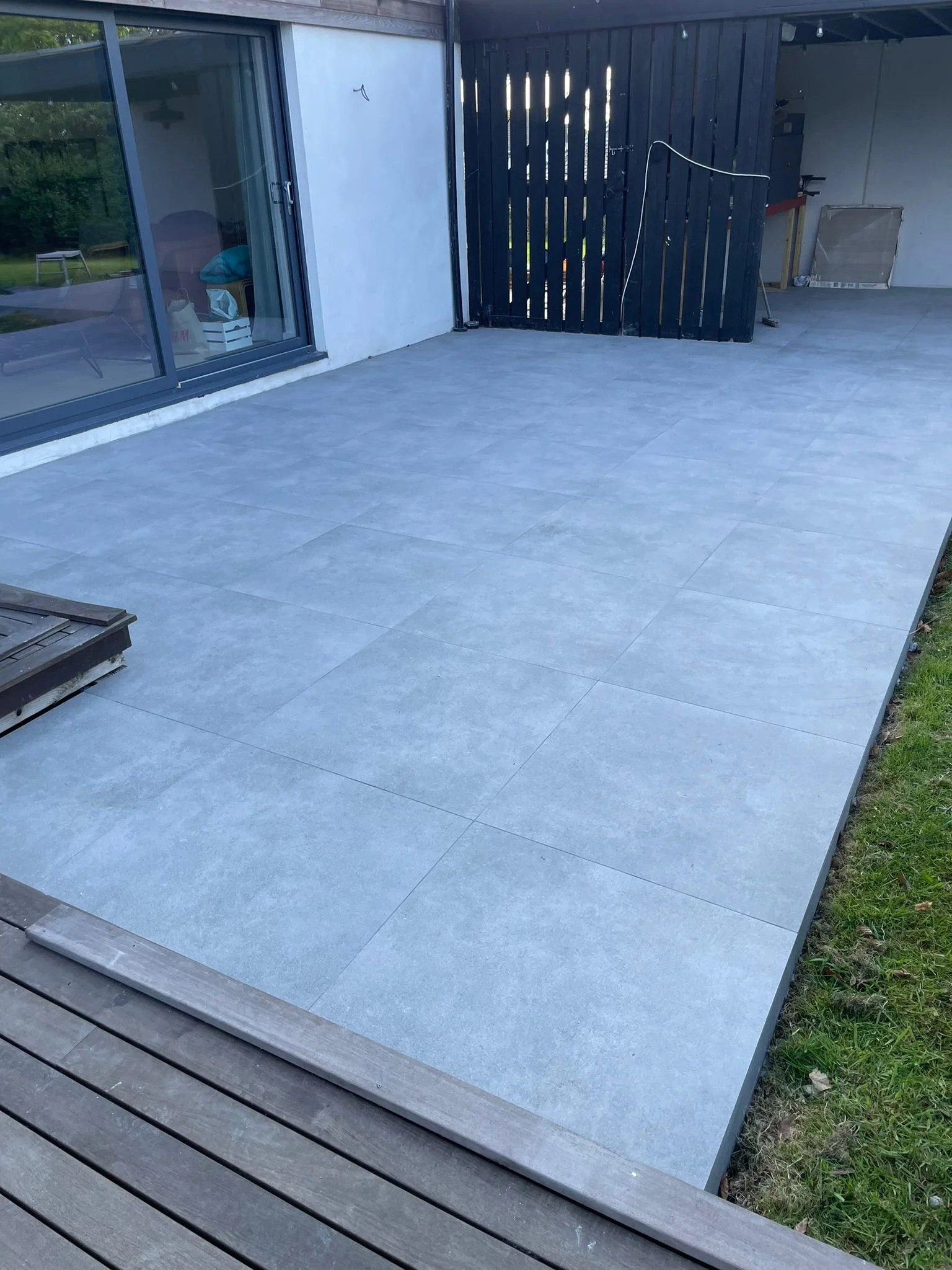 A newly finished outdoor patio with gray tiles next to a house, with a black wooden fence and a sliding glass door.