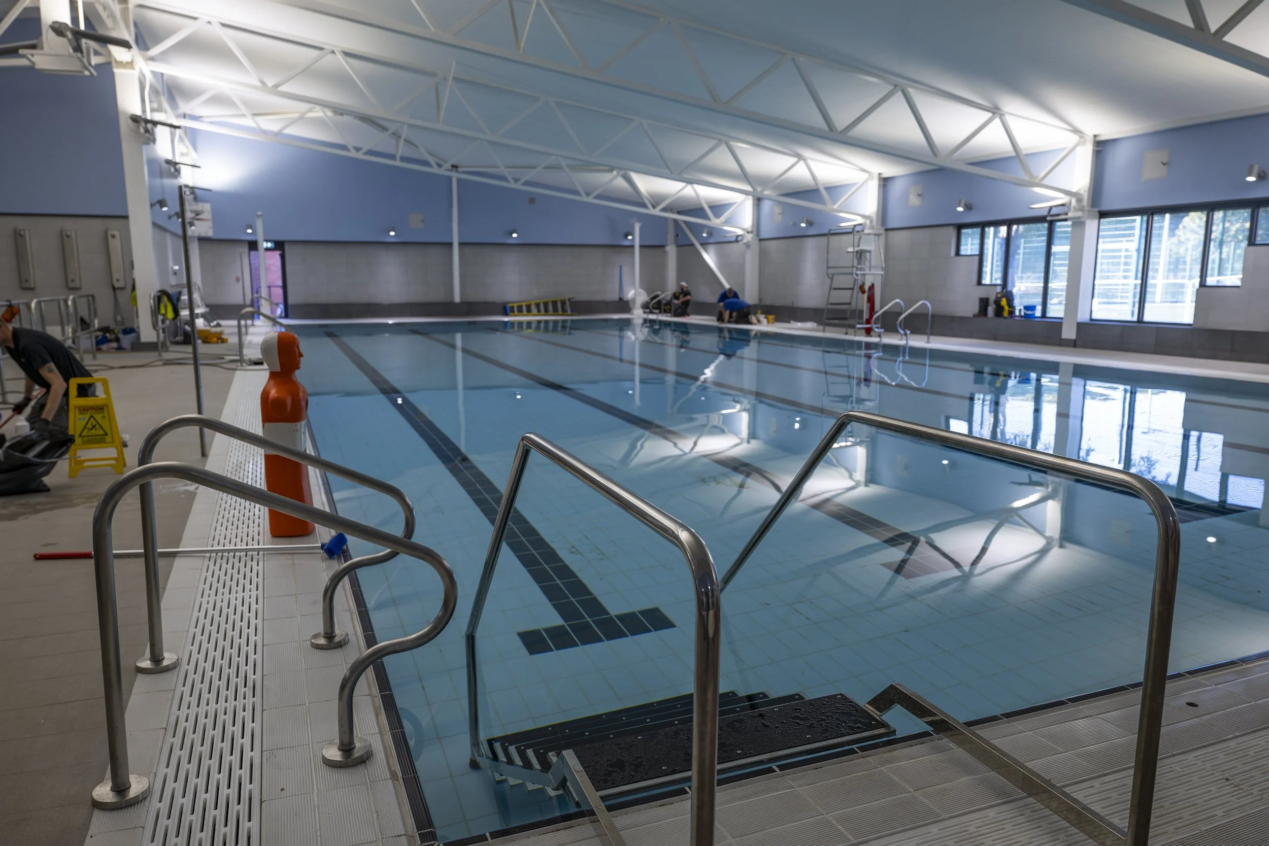 Empty indoor swimming pool with metal handrails, pool cleaning equipment, and workers in the background cleaning the pool.