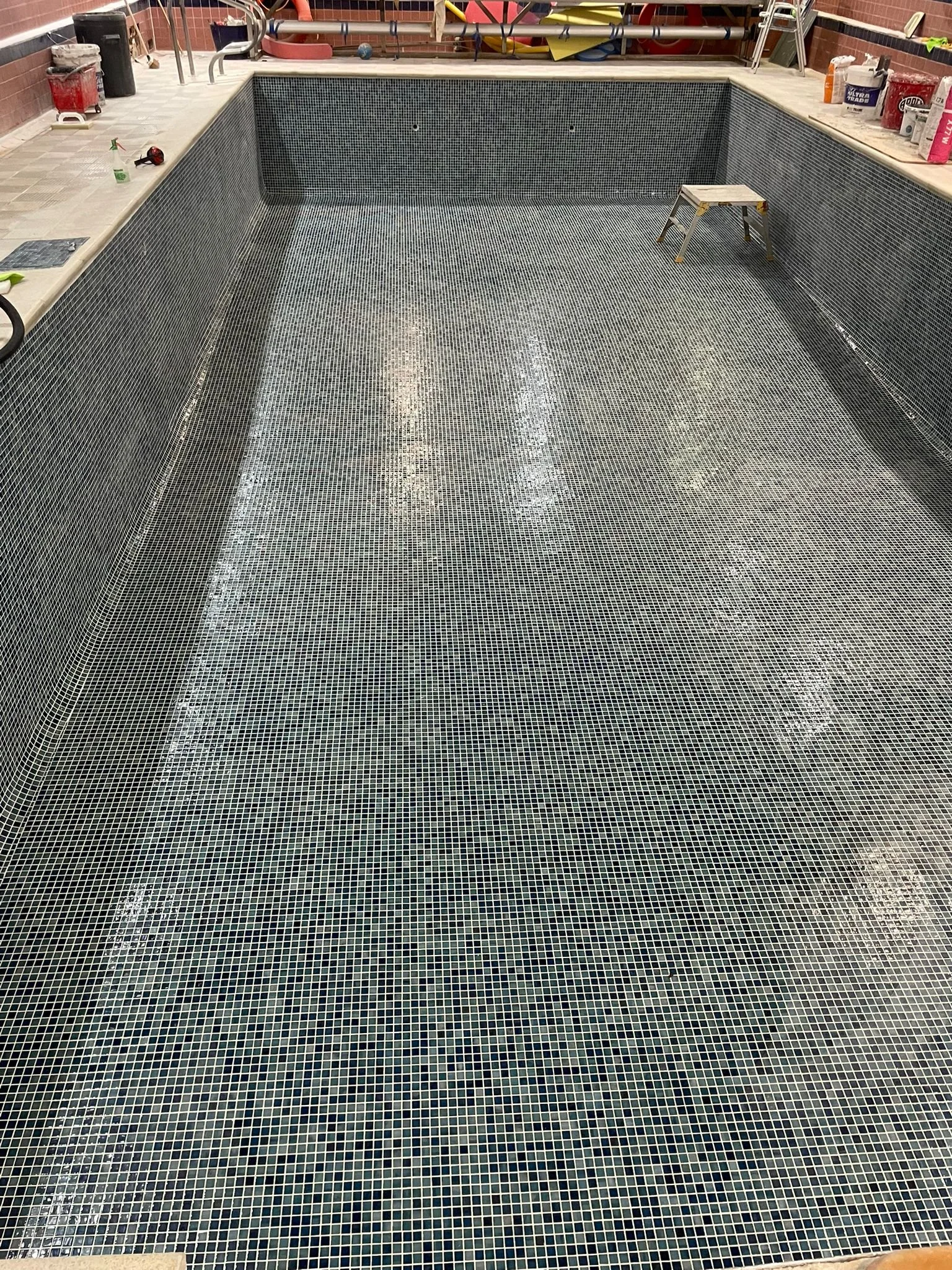 Empty swimming pool with mosiac tile lining, construction tools and materials around the edges, and a small step ladder inside.