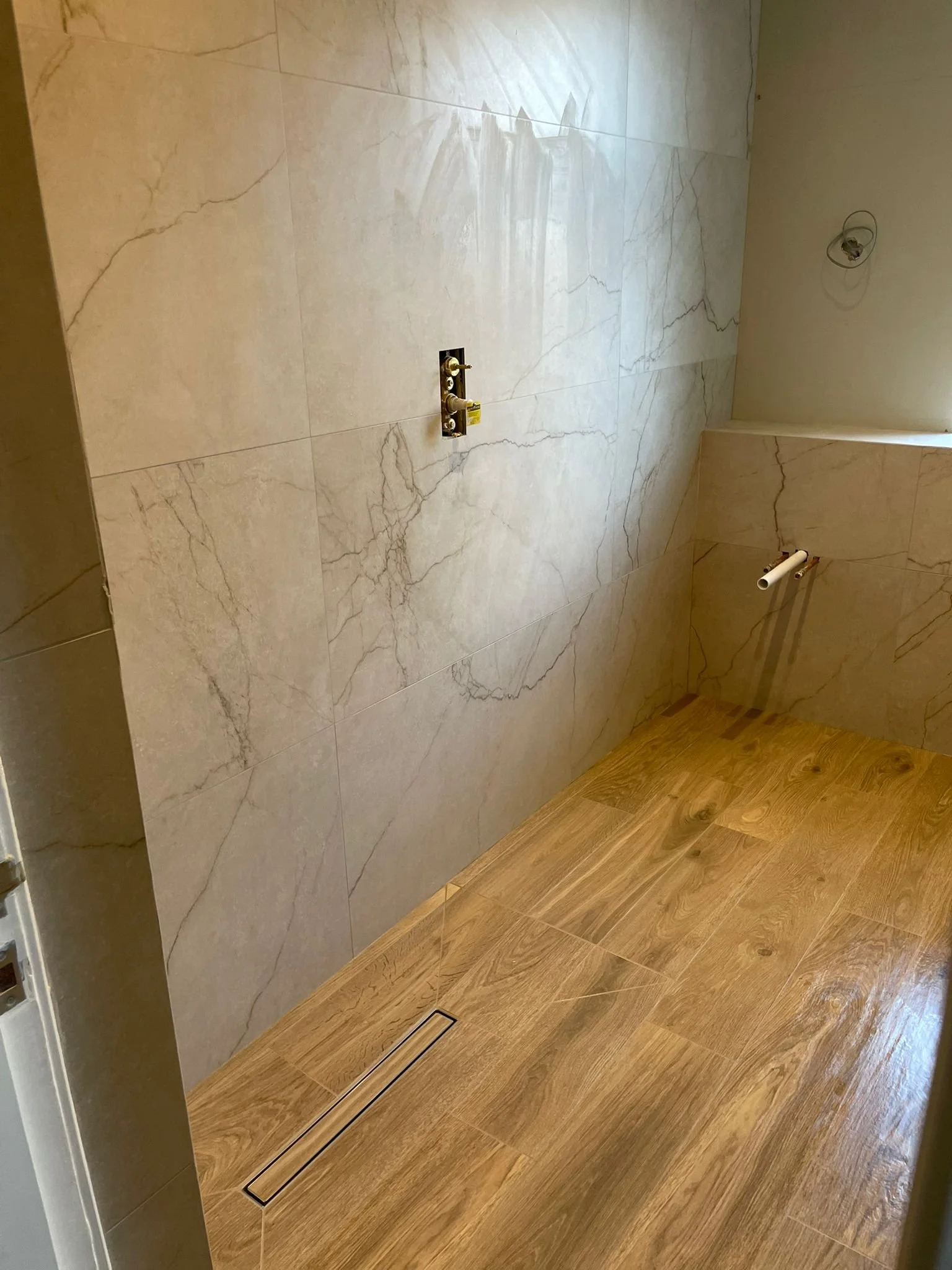 A partially finished bathroom with marble wall tiles, wooden floor, plumbing fixtures, and an electrical box on the wall.