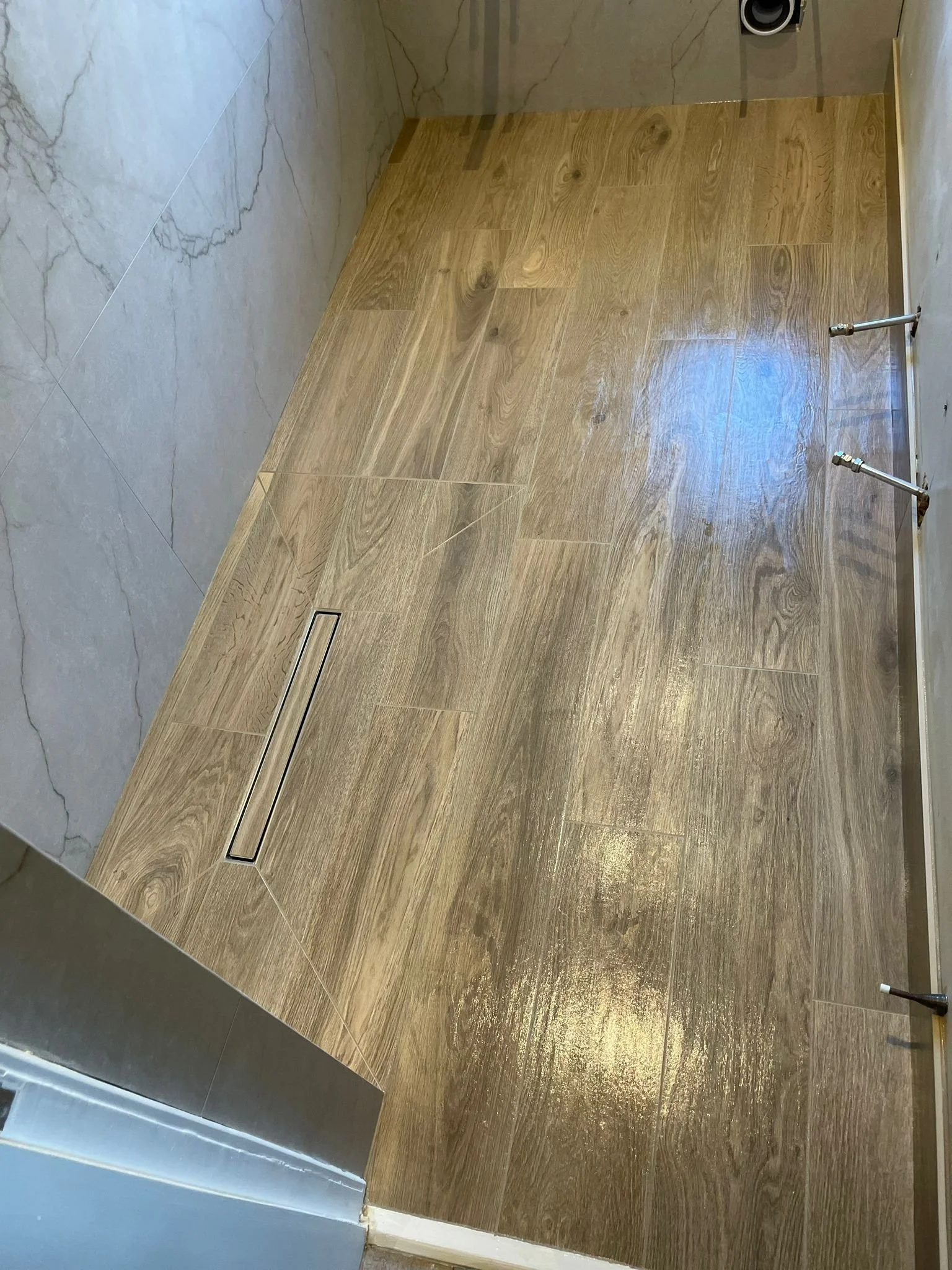 Bathroom floor with light wood and marble tiles, drain, and plumbing pipes.