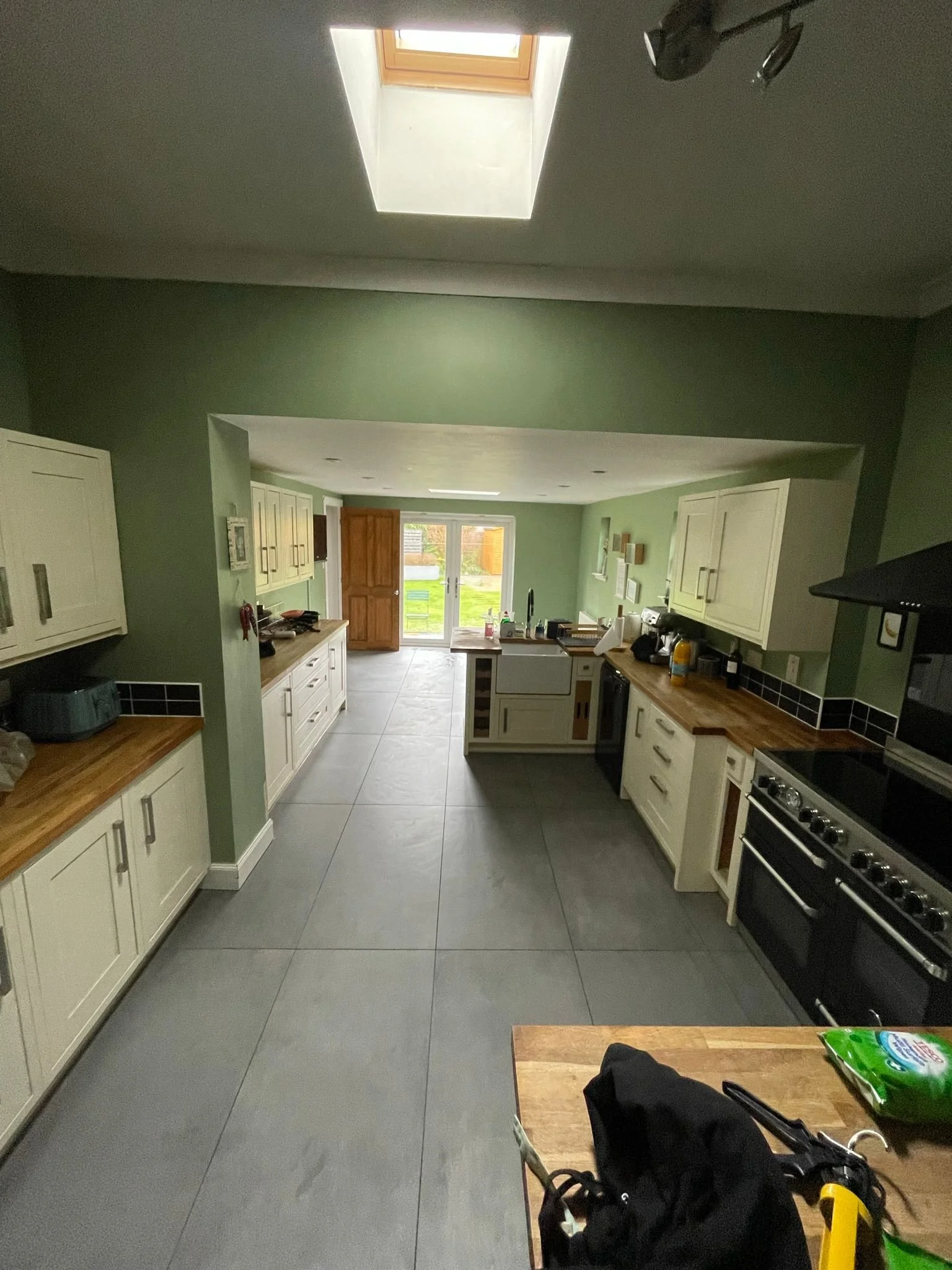 A kitchen with light green walls, white cabinetry with wooden countertops, gray floor tiles, and a skylight in the ceiling. There are double doors to the outside, kitchen appliances, and a wooden table with items on it in the foreground.