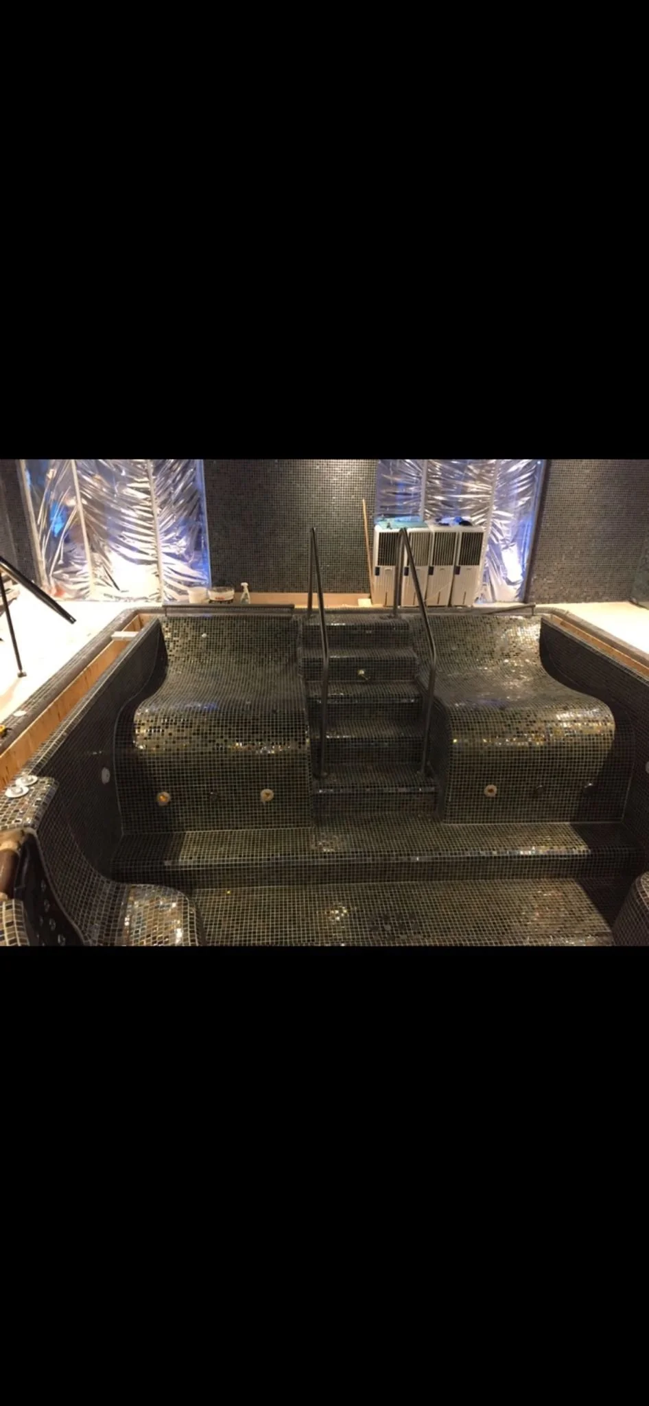 Indoor hot tub or jacuzzi with black mosaic tiles, black metal handrails, and steps leading into the water, surrounded by a modern decorative wall with metallic patterns.