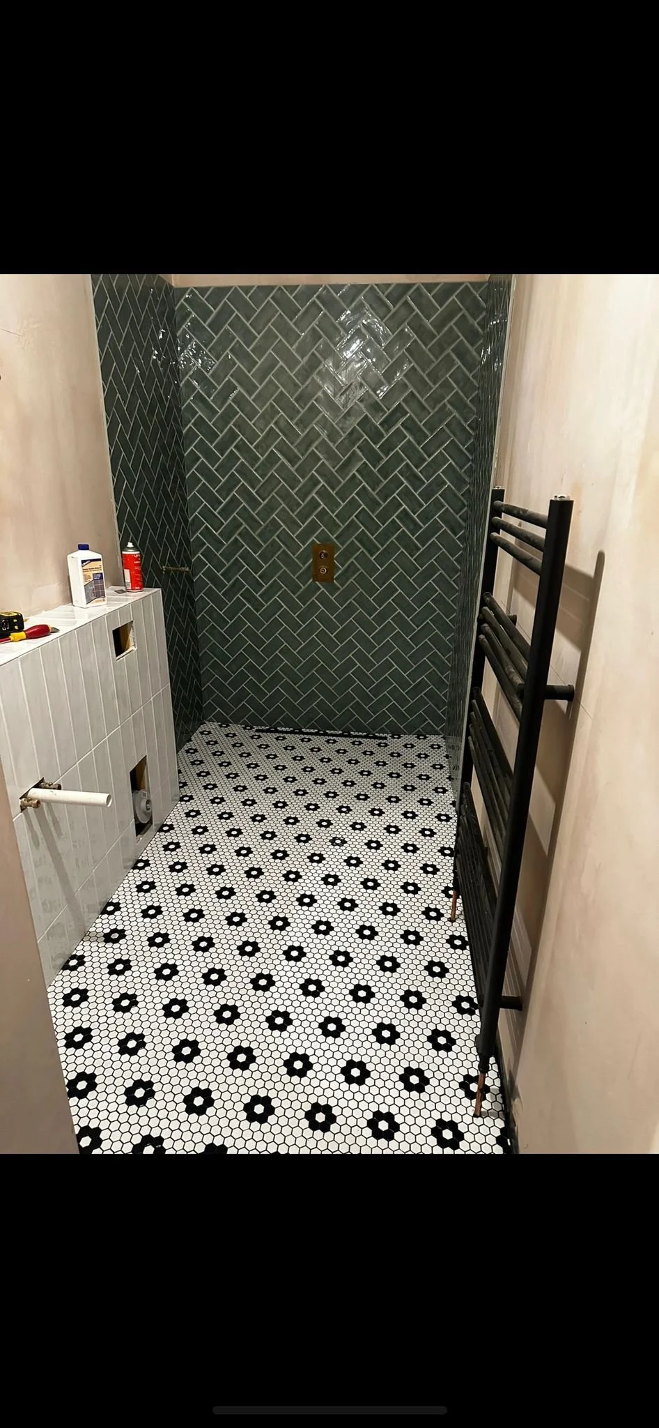 A bathroom under renovation with black and white hexagonal patterned floor tiles, a dark herringbone tiled wall in the shower area, and building tools and supplies on a partially tiled wall.