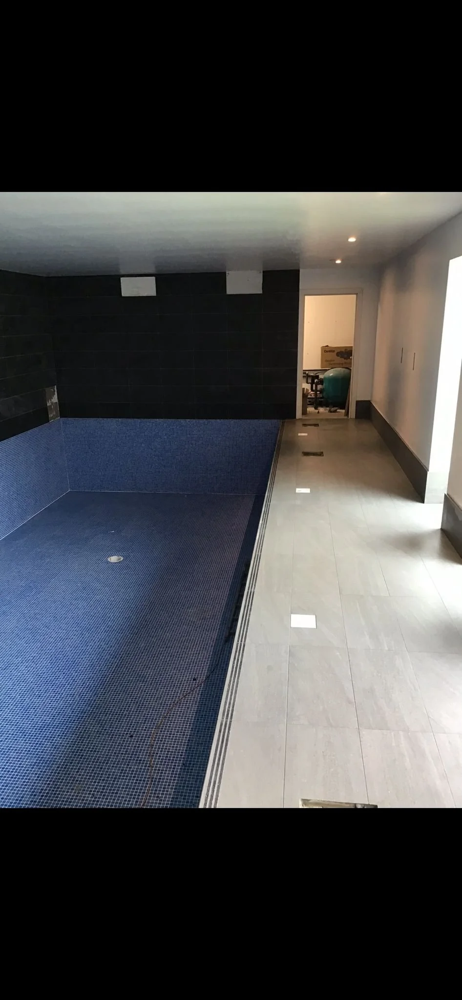Indoor pool area with blue mosaic tile floor and black tiled wall, next to a hallway leading to a room with gym equipment in the background.
