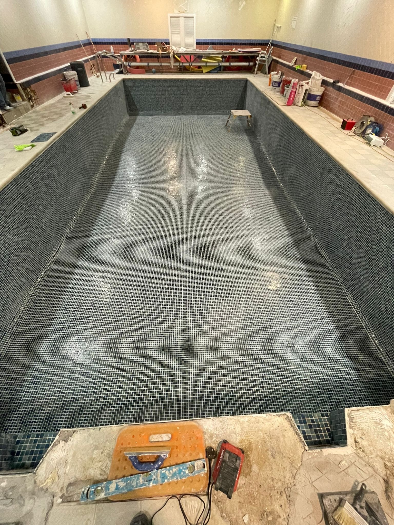 Indoor swimming pool under maintenance with tools and supplies, empty and tiled with small black tiles.