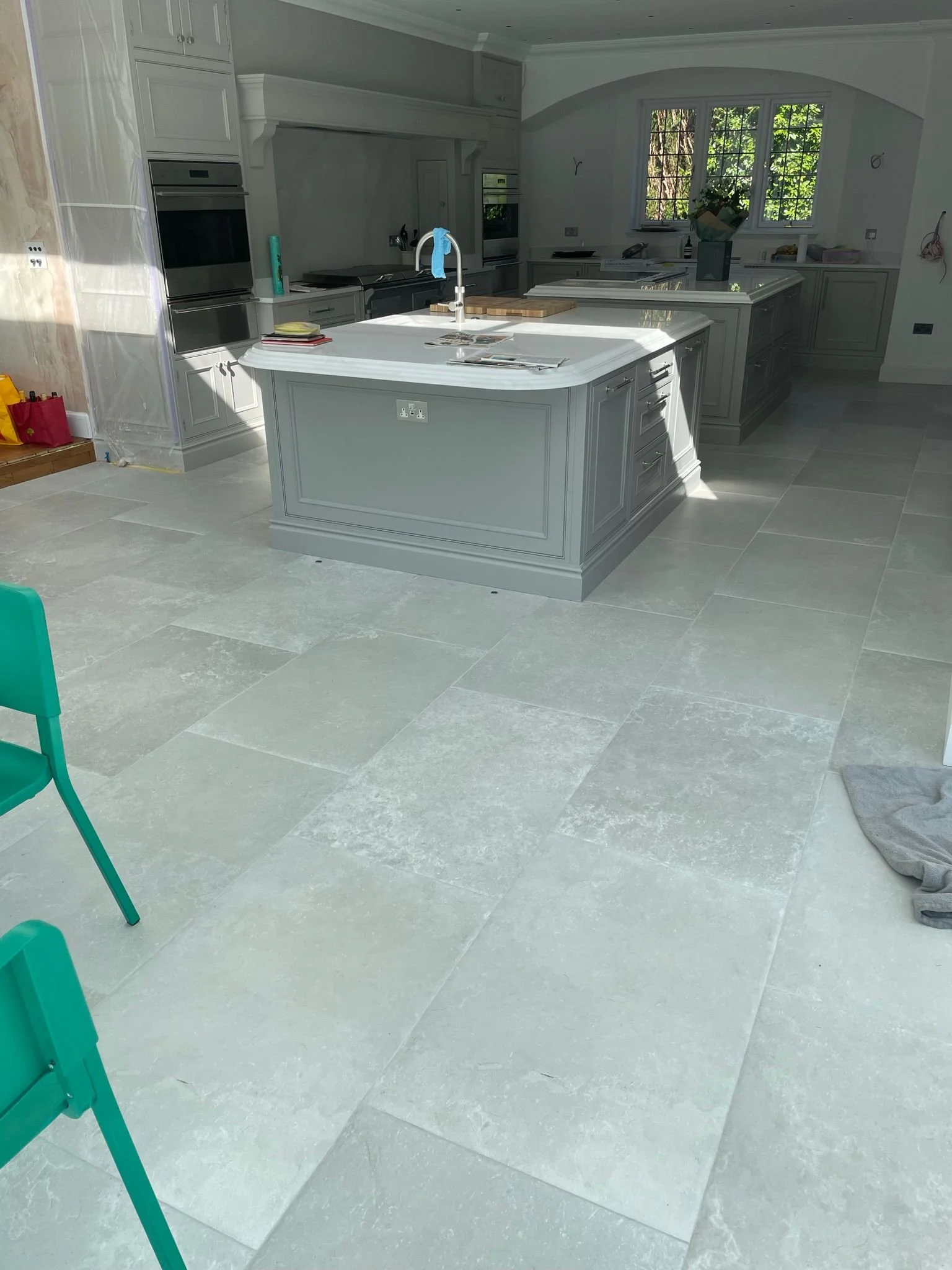 Modern kitchen with large central island, built-in appliances, light gray cabinetry, large tiled floor, and a window view of greenery outside.