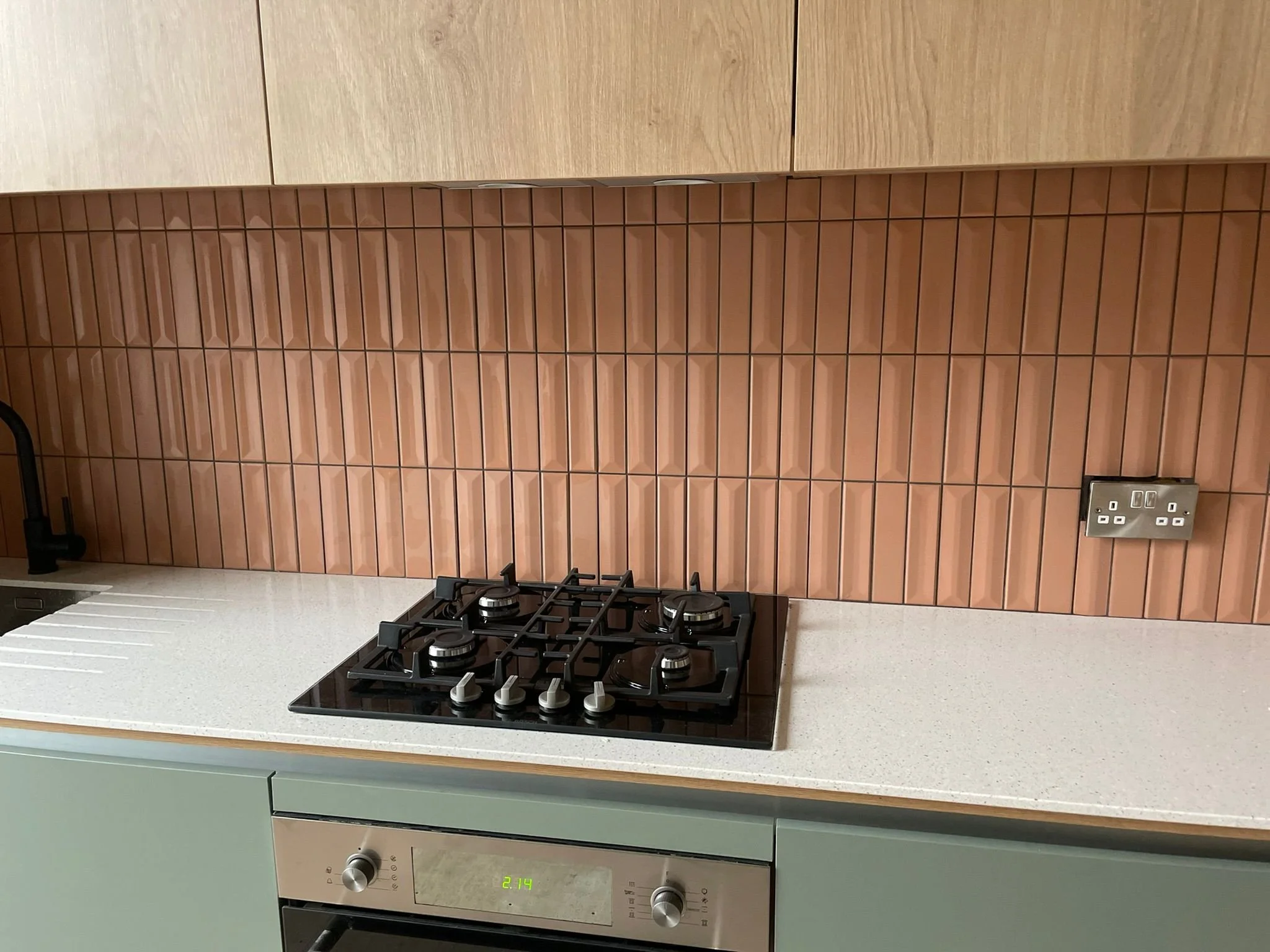 Modern kitchen countertop with built-in gas stove, light-colored speckled surface, pink tiled backsplash, and light wood upper cabinets.