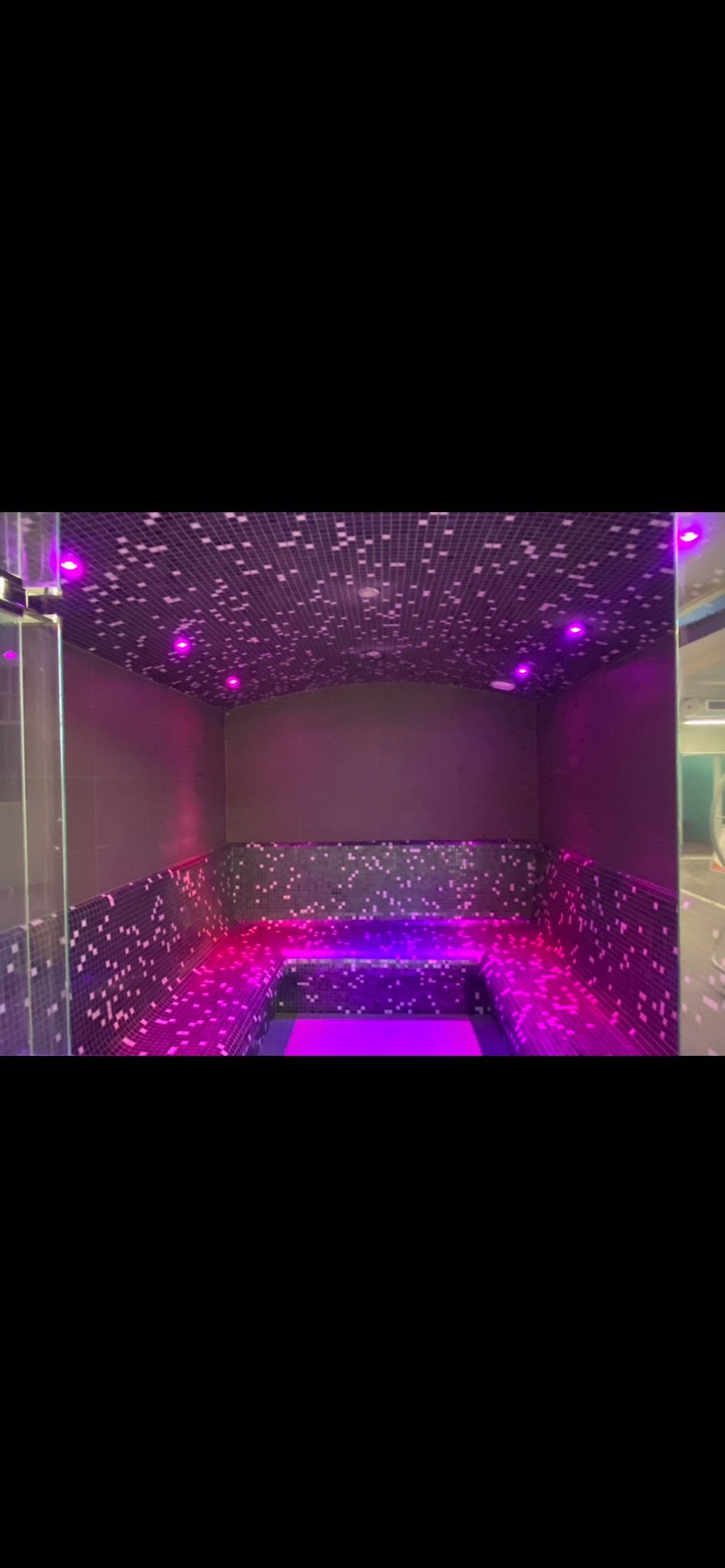 A spa or hot tub area with black mosaic tile design and purple LED lighting, featuring a curved seating area and a tiled ceiling with small square patterns.